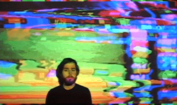 A person with dark hair and a beard standing in front of a colorful, distorted background that resembles a digital glitch or screen error.