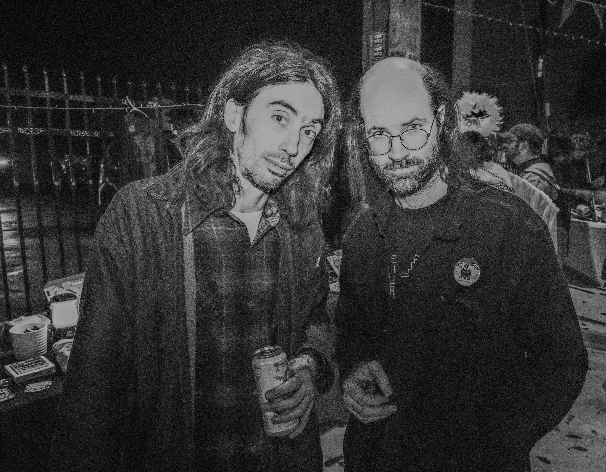 Two men with long hair and beards standing close together outdoors at night, one holding a can of drink, with a fence and other people in the background.
