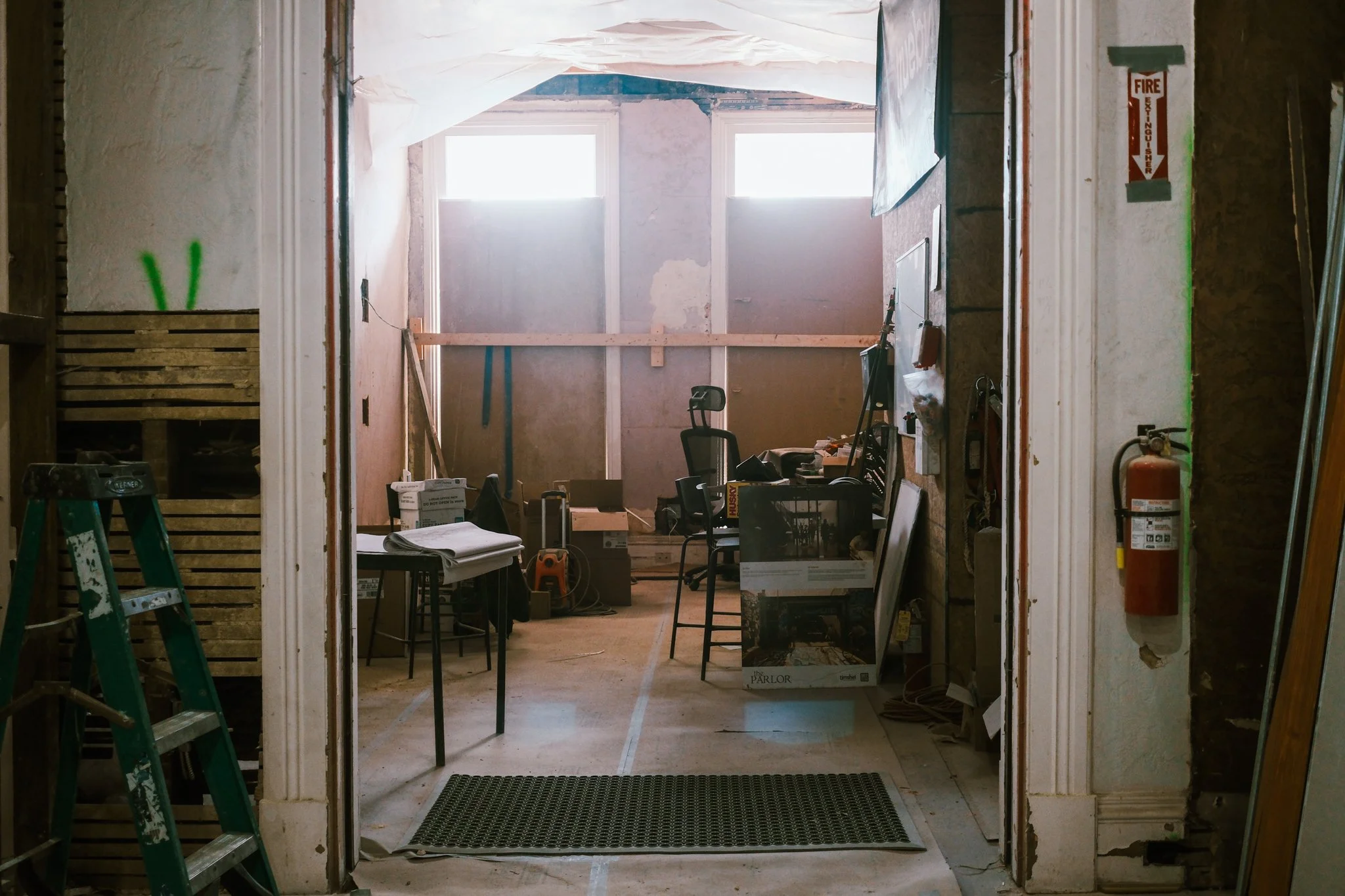 Interior of a construction site with tools, furniture, and building materials. Wood planks, a green ladder, and a fire extinguisher are visible. The space is unfinished with exposed walls and large windows letting in natural light.