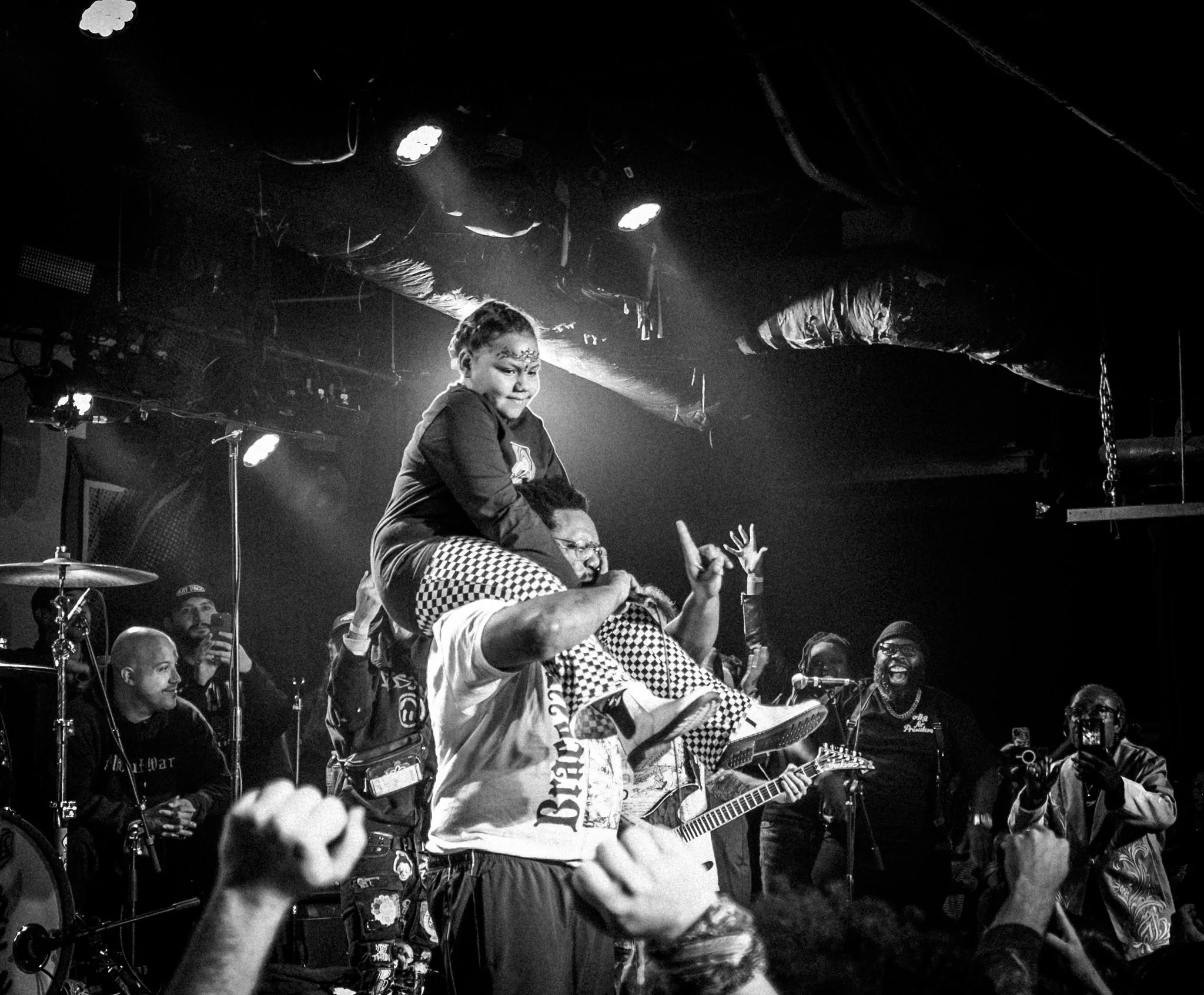 A lively concert scene in black and white with a group of musicians and fans. A woman is sitting on a man's shoulders, raising her hand while singing or cheering. Several people are smiling, playing instruments, or capturing the moment on their phone