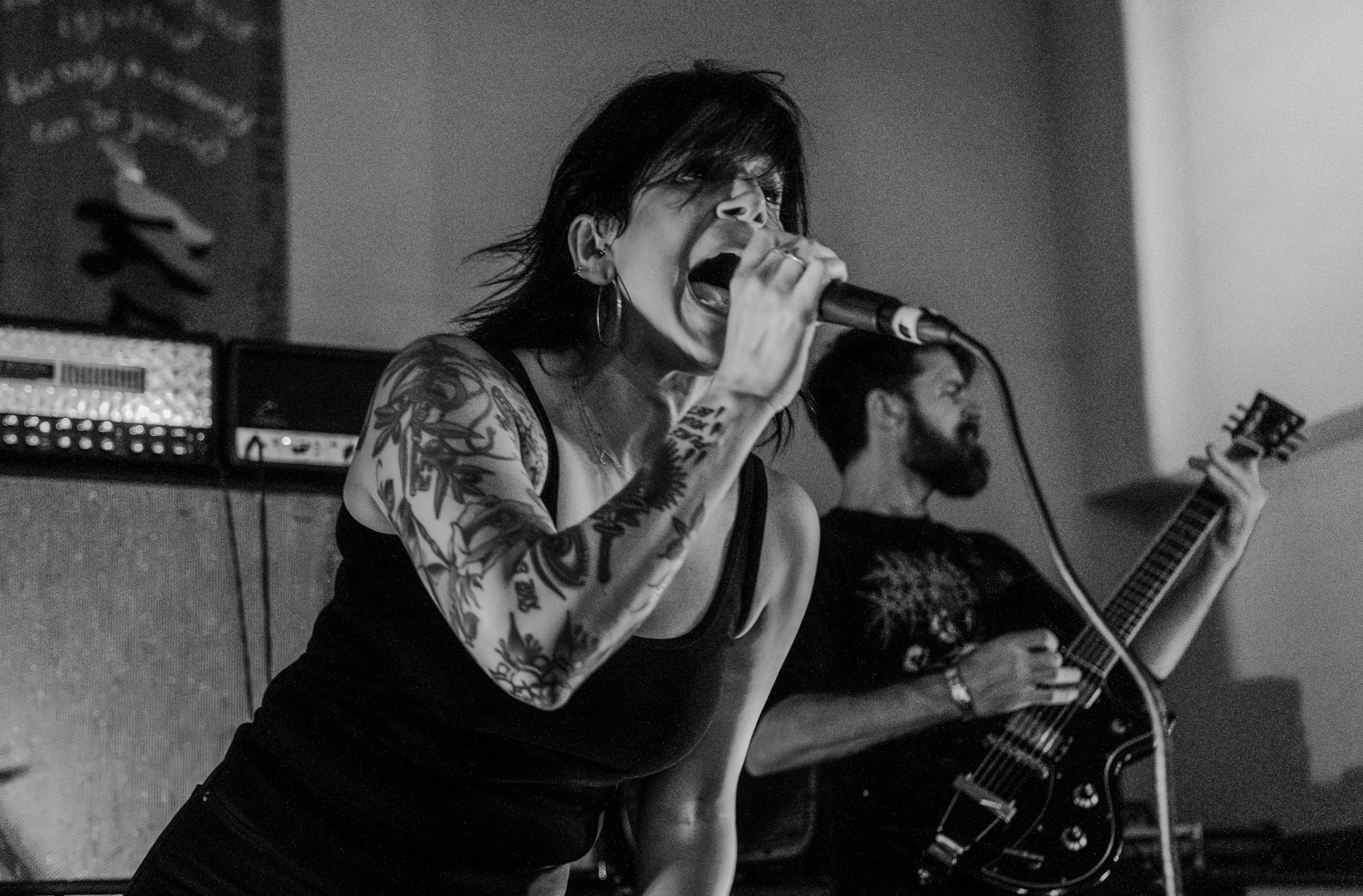 A female vocalist with tattoos singing into a microphone during a live music performance, with a guitarist playing guitar in the background.