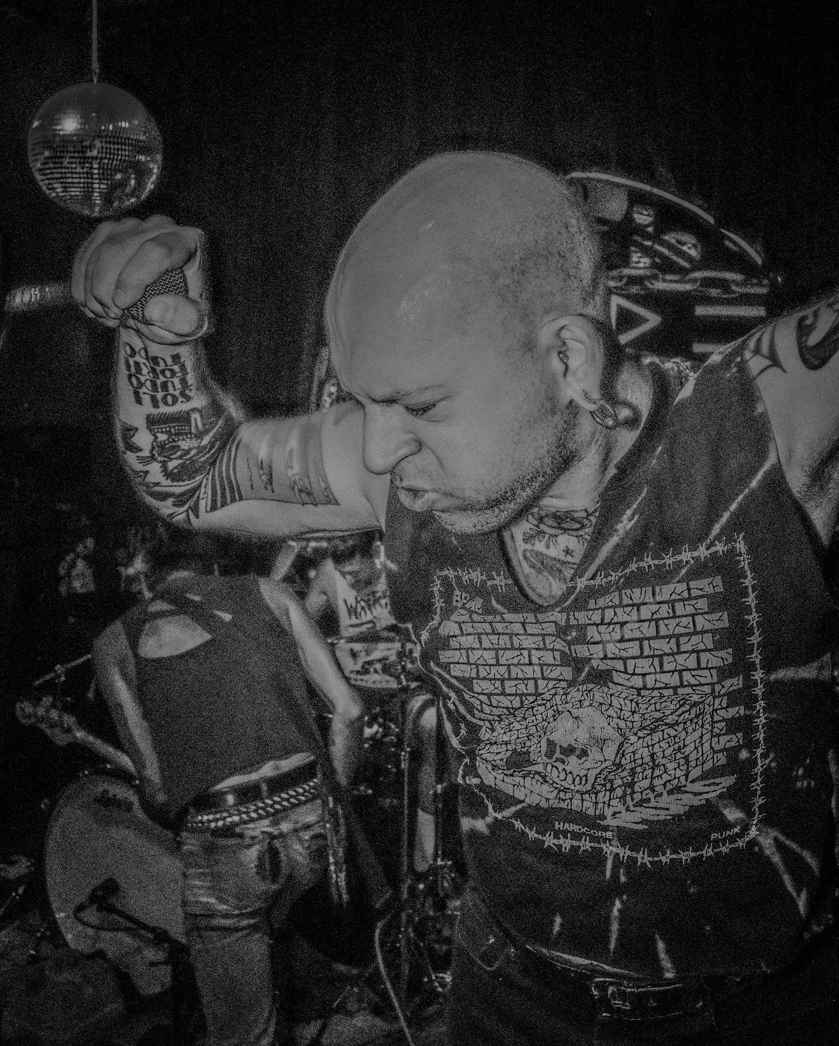 A punk rock musician with tattoos on his arm and chest, wearing a black sleeveless shirt with a skull graphic, performing passionately in a dimly lit setting with a disco ball overhead.