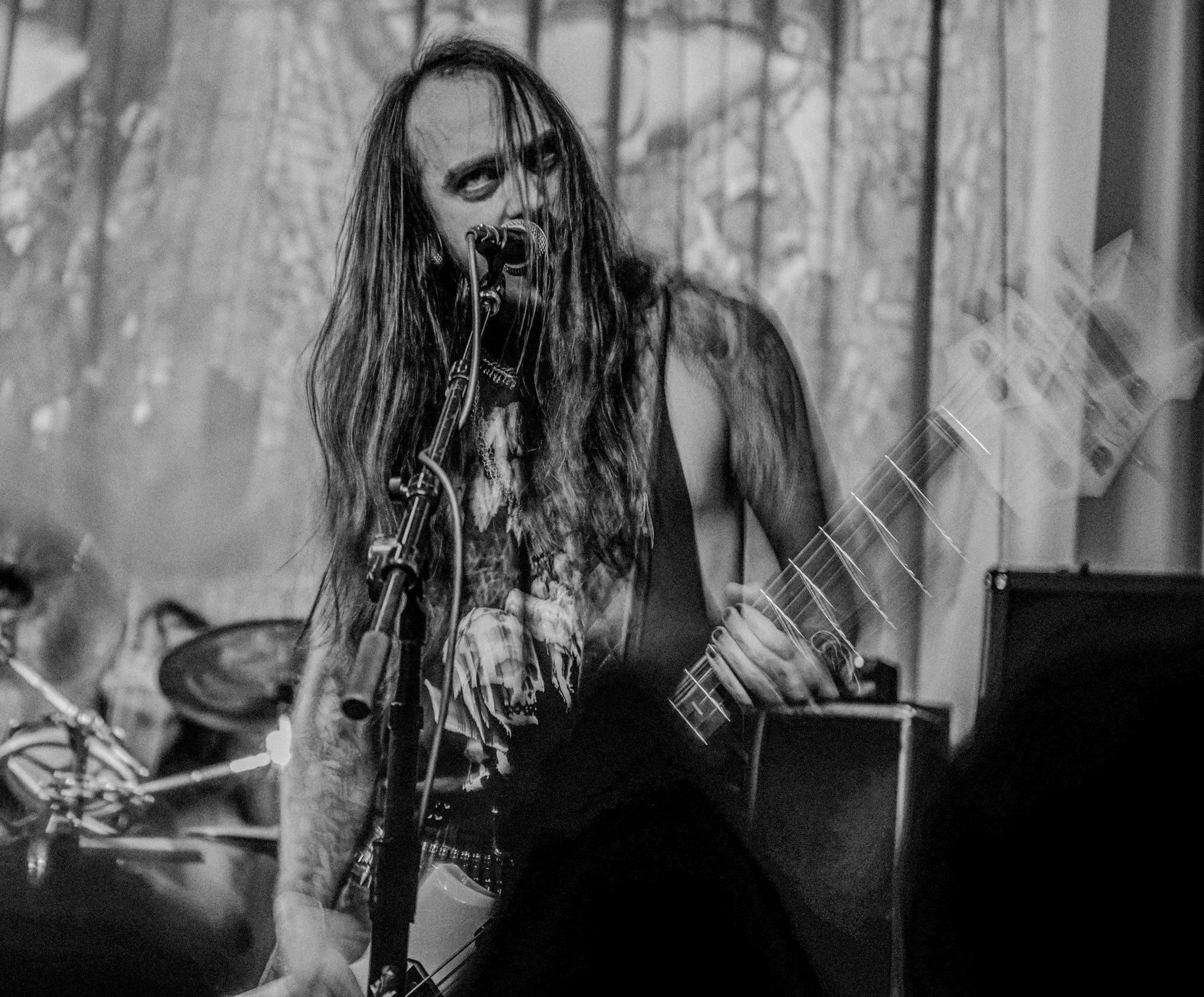 A female musician with long hair, tattoos, and goth-style makeup singing into a microphone while playing an electric guitar during a live performance.
