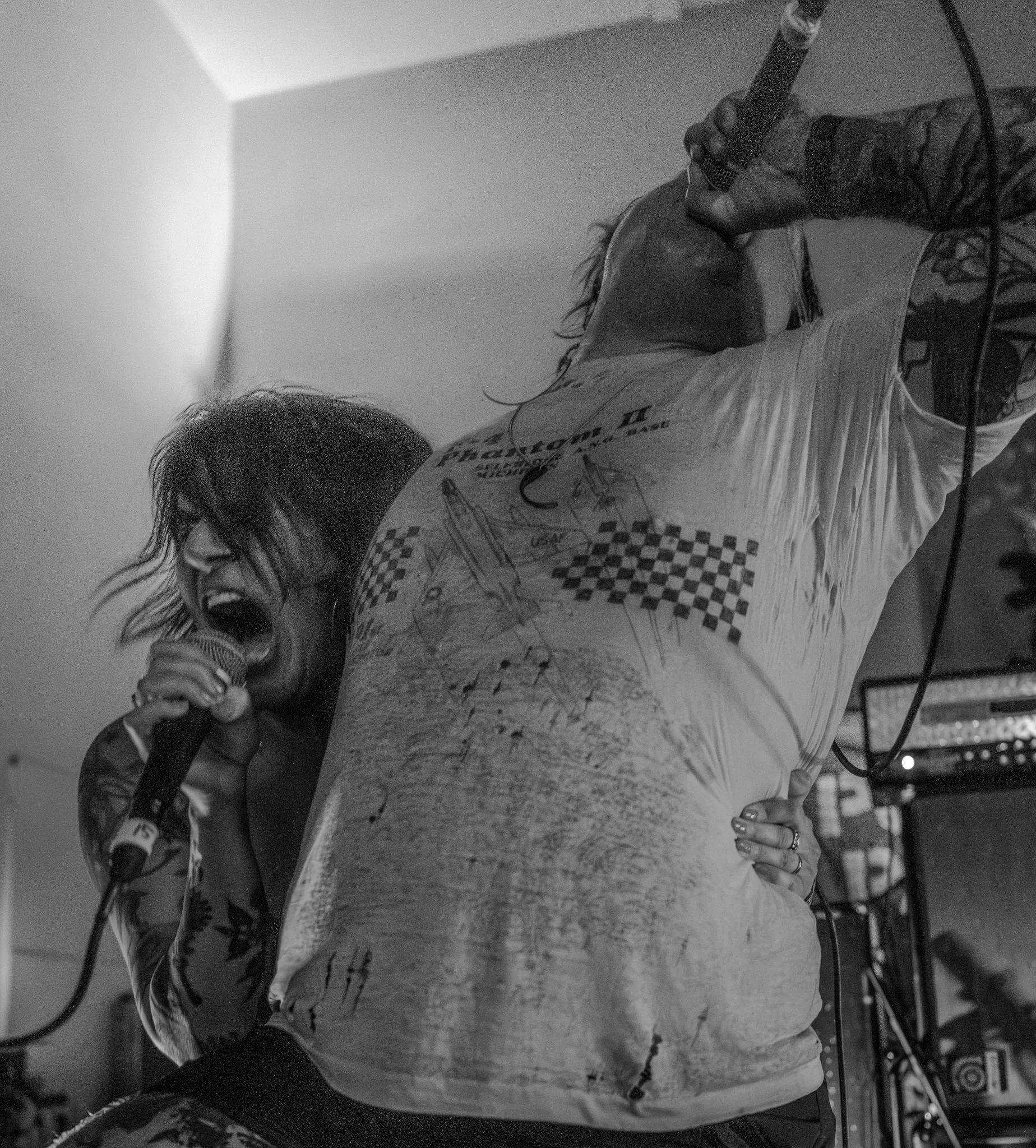 A black and white photo of two musicians performing passionately on stage. One is singing into a microphone with an intense expression, and the other is holding a microphone close to their mouth. The person with the microphone has tattoos on their ar
