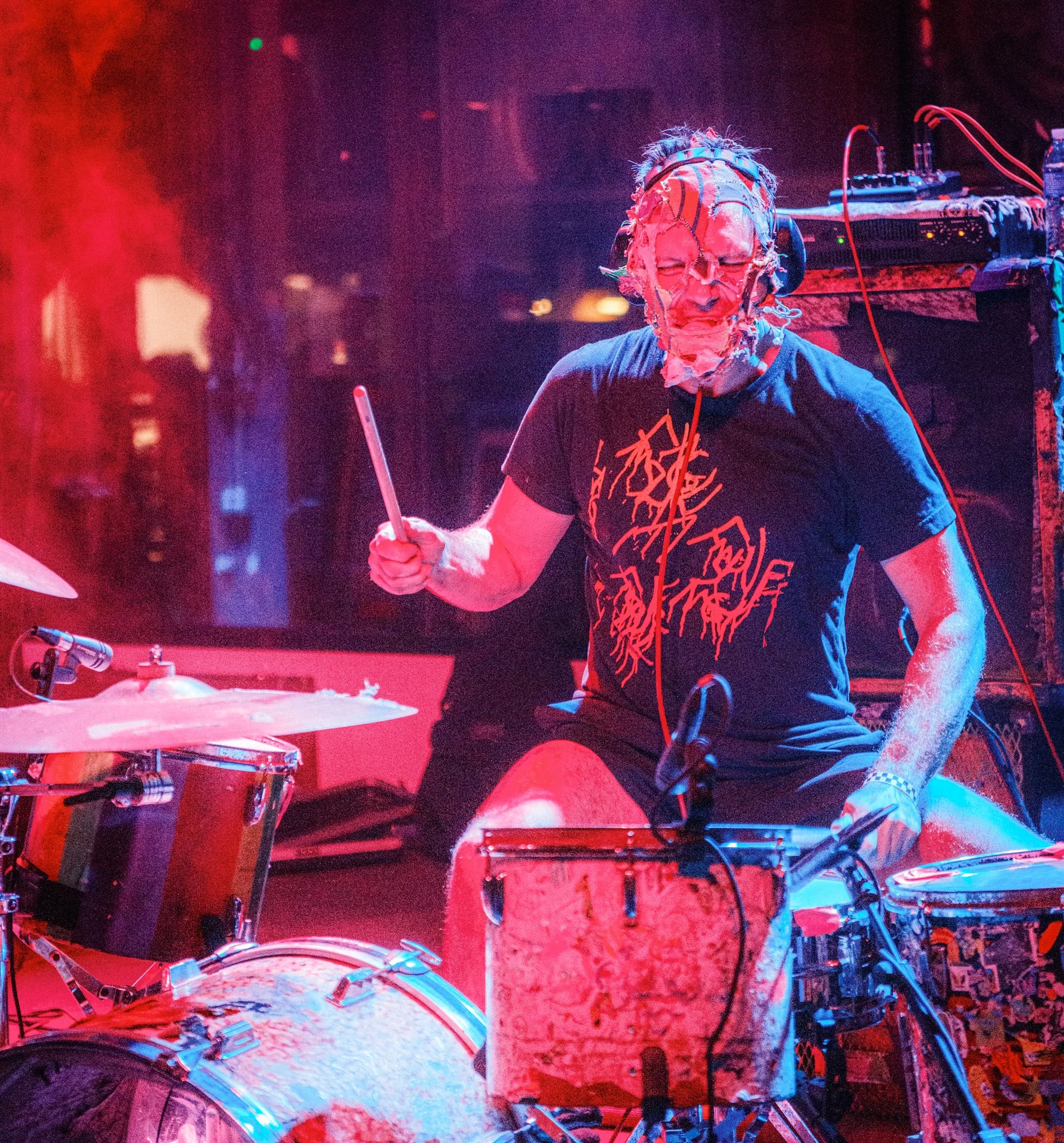 A drummer wearing a mask with wires and tattered fabric, playing on a drum set in a dimly lit, colorful concert stage environment.