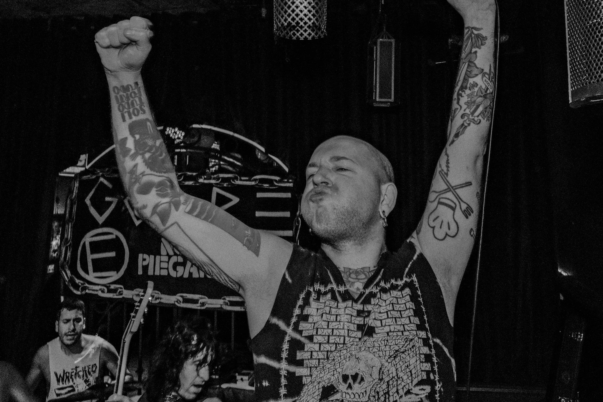 A man with tattoos on his arms and earrings, wearing a sleeveless shirt with a skull design, is raising his fist in the air on stage at a concert or performance.