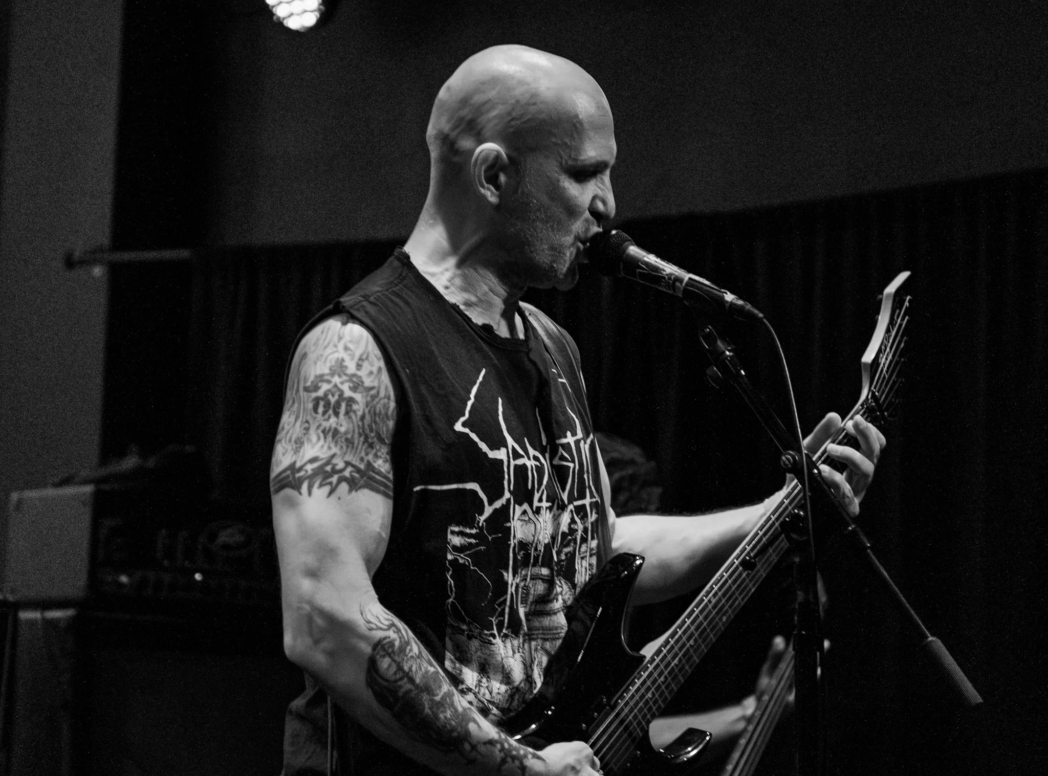 A tattooed man with a shaved head singing into a microphone while playing an electric guitar in a dark room.