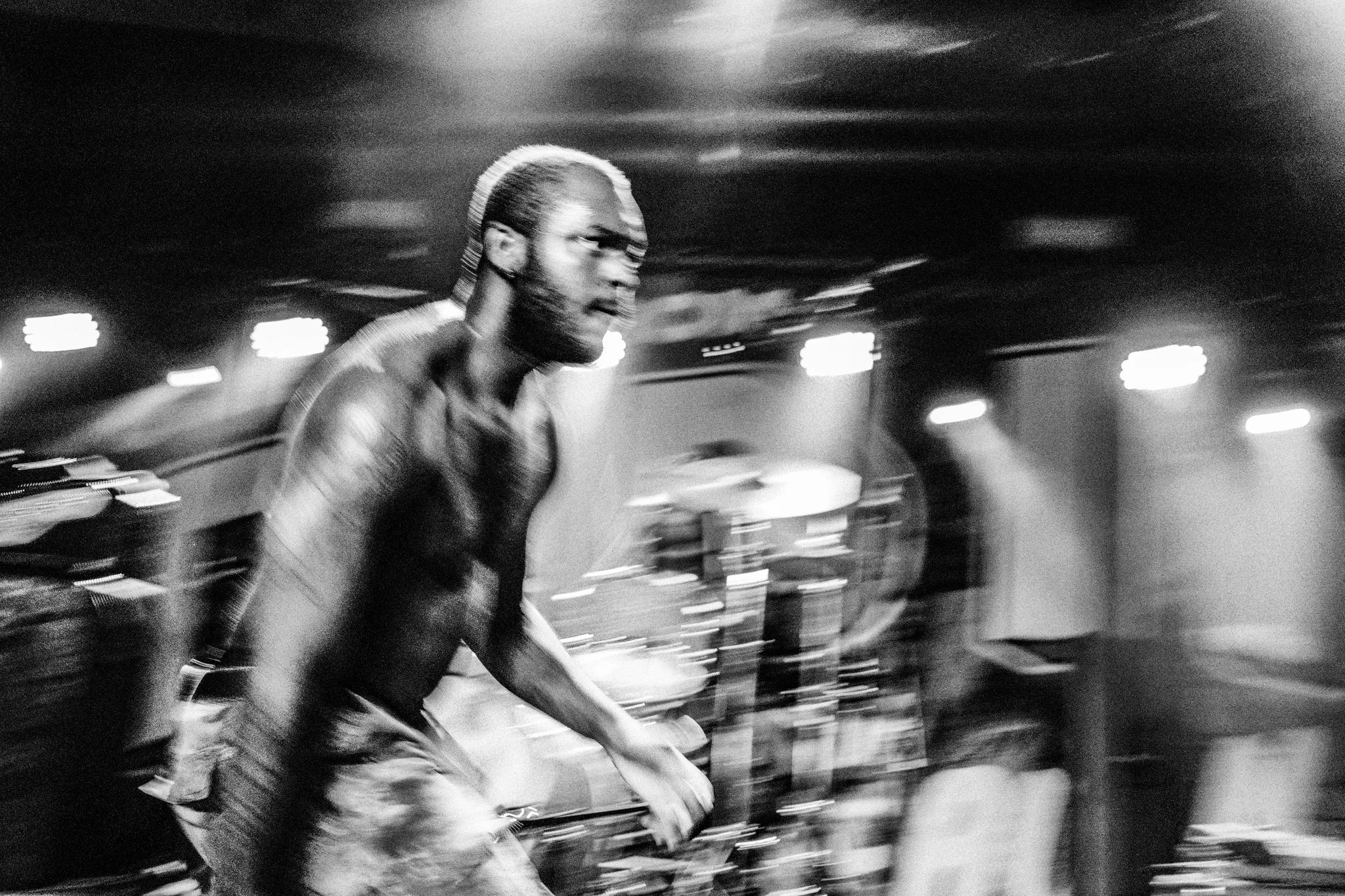 A black and white photo of a shirtless man with glasses and a beard, appearing to be in motion, possibly performing or dancing in a dimly lit room with stage lights.