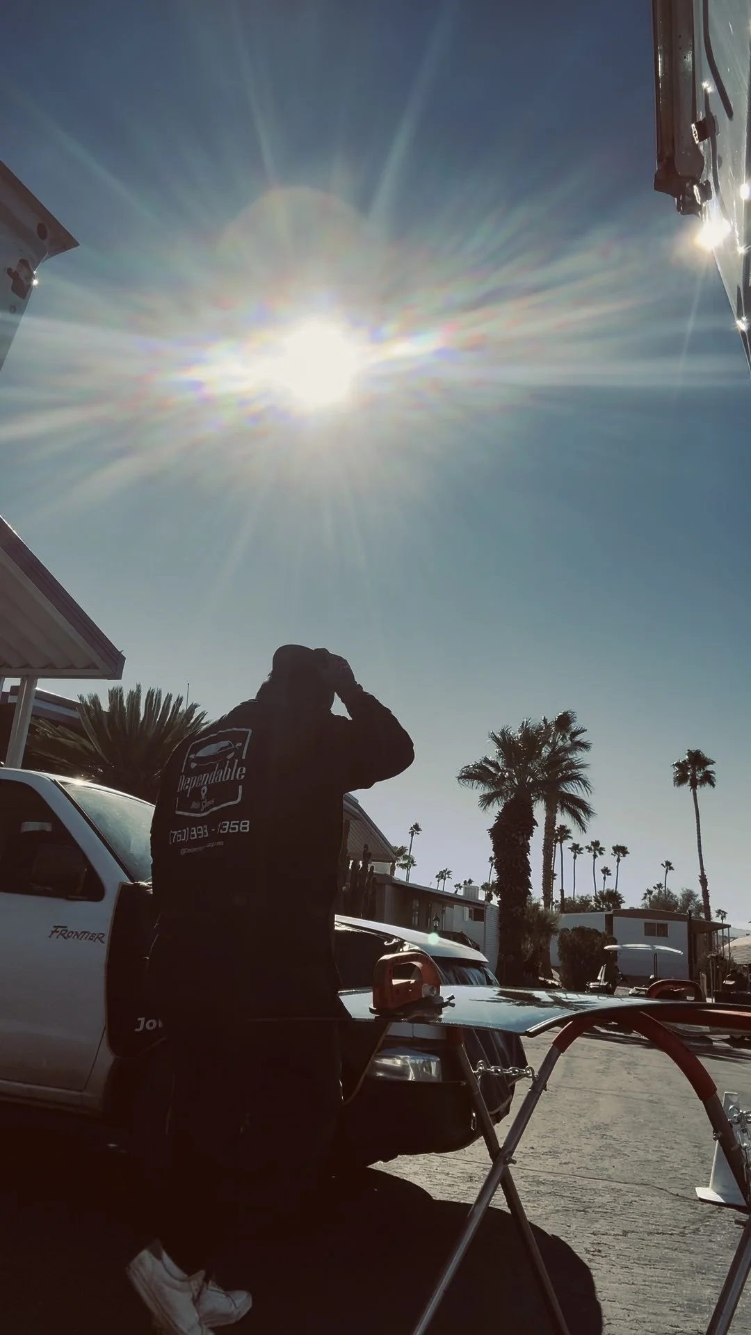 A person standing next to a white pickup truck, with tall palm trees in the background, under a bright, sunny sky with a lens flare effect.