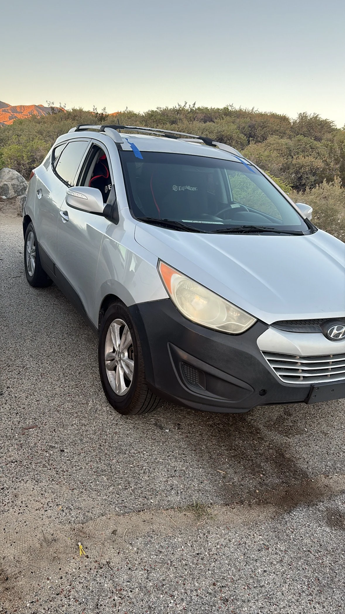 A silver Hyundai hatchback is parked on a gravel surface with a desert landscape and mountains in the background, under a clear sky.