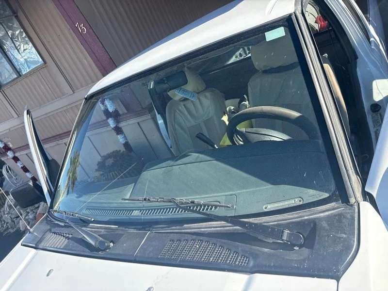 Front view of a white vehicle, showing the windshield, steering wheel, and front seats. The vehicle is parked near a building with maroon and beige siding.
