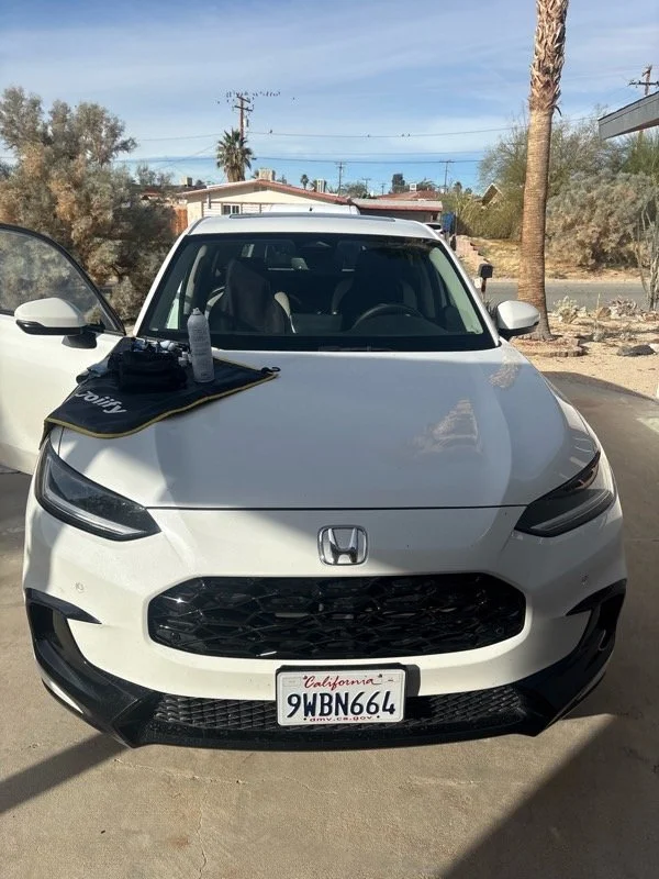 Front view of a white Honda car parked outdoors during the daytime, with a license plate reading California 9WBN664, a palm tree in the background, and a black pouch or bag and a water bottle placed on the hood.