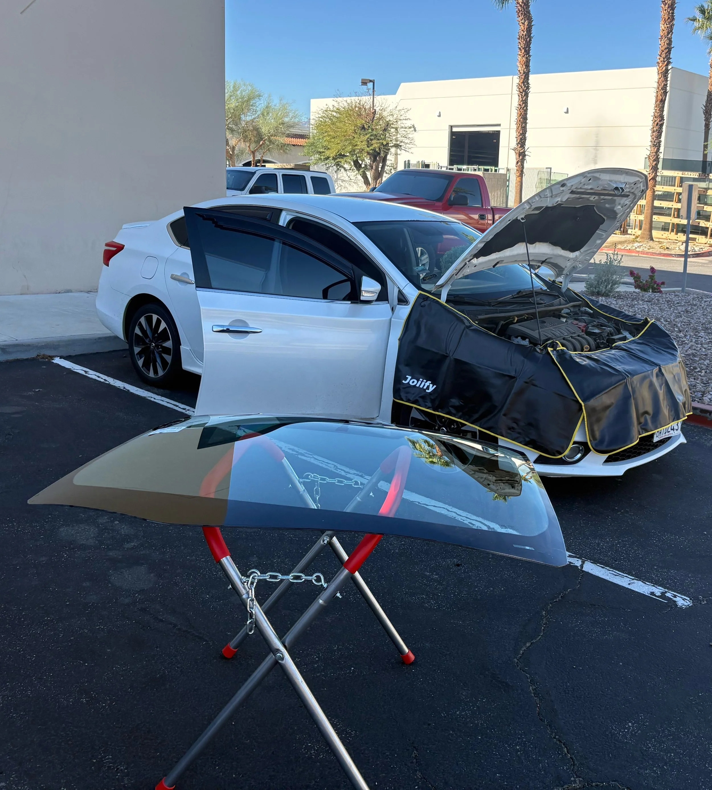 A white sedan car with its hood open and a black cover over the front end, parked in an outdoor parking lot next to a white building. In front of the car, there is a small collapsible table with a black carbon fiber hood resting on it.