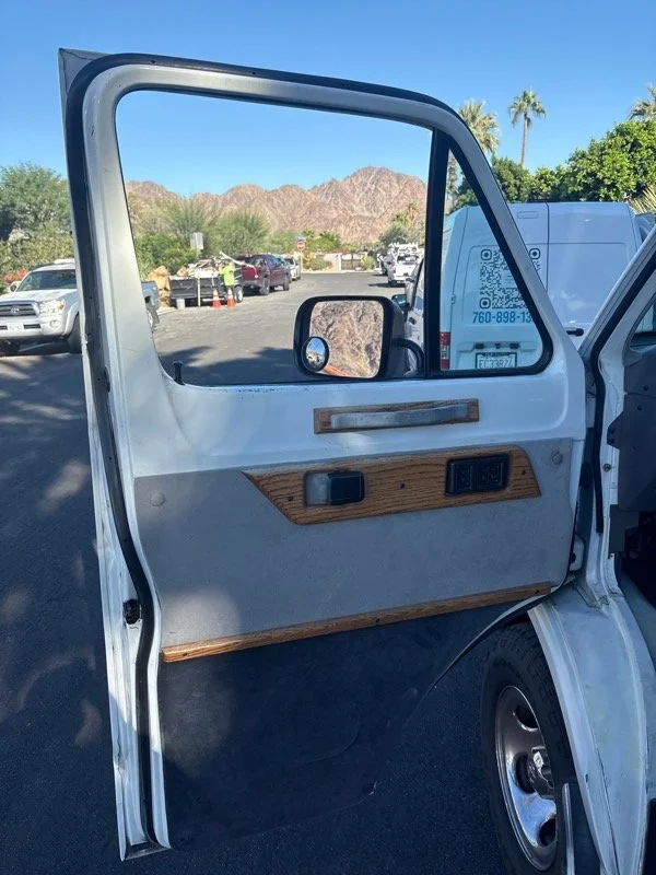 Open vehicle door with brown wood trim, vehicle locking controls, and side mirror, with a parking lot, trees, mountains, and clear blue sky in background.