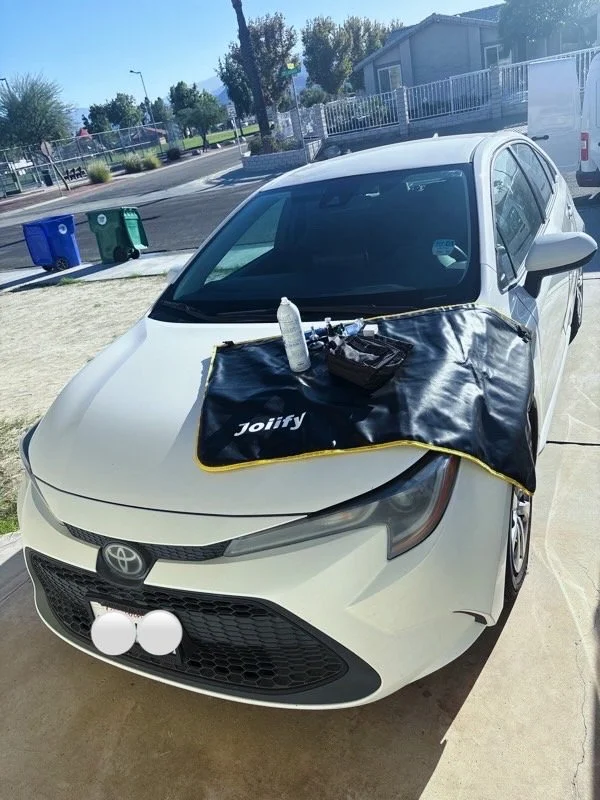White Toyota car parked outdoors with a black padded mat labeled 'Joify' on the hood, holding cleaning supplies including spray bottles and cleaning cloths.