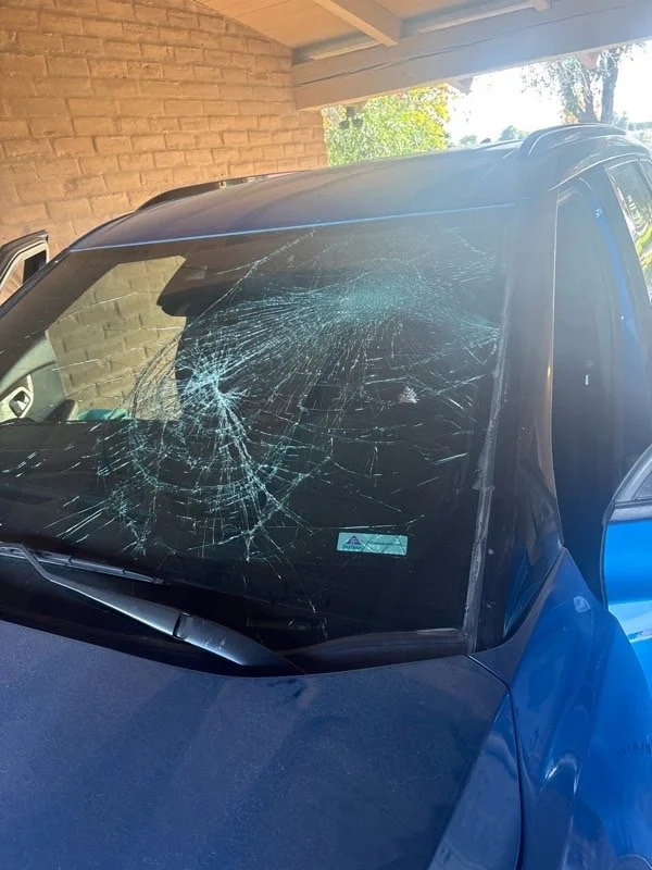 Blue vehicle with heavily cracked windshield under a carport with brick wall and tree visible in the background.