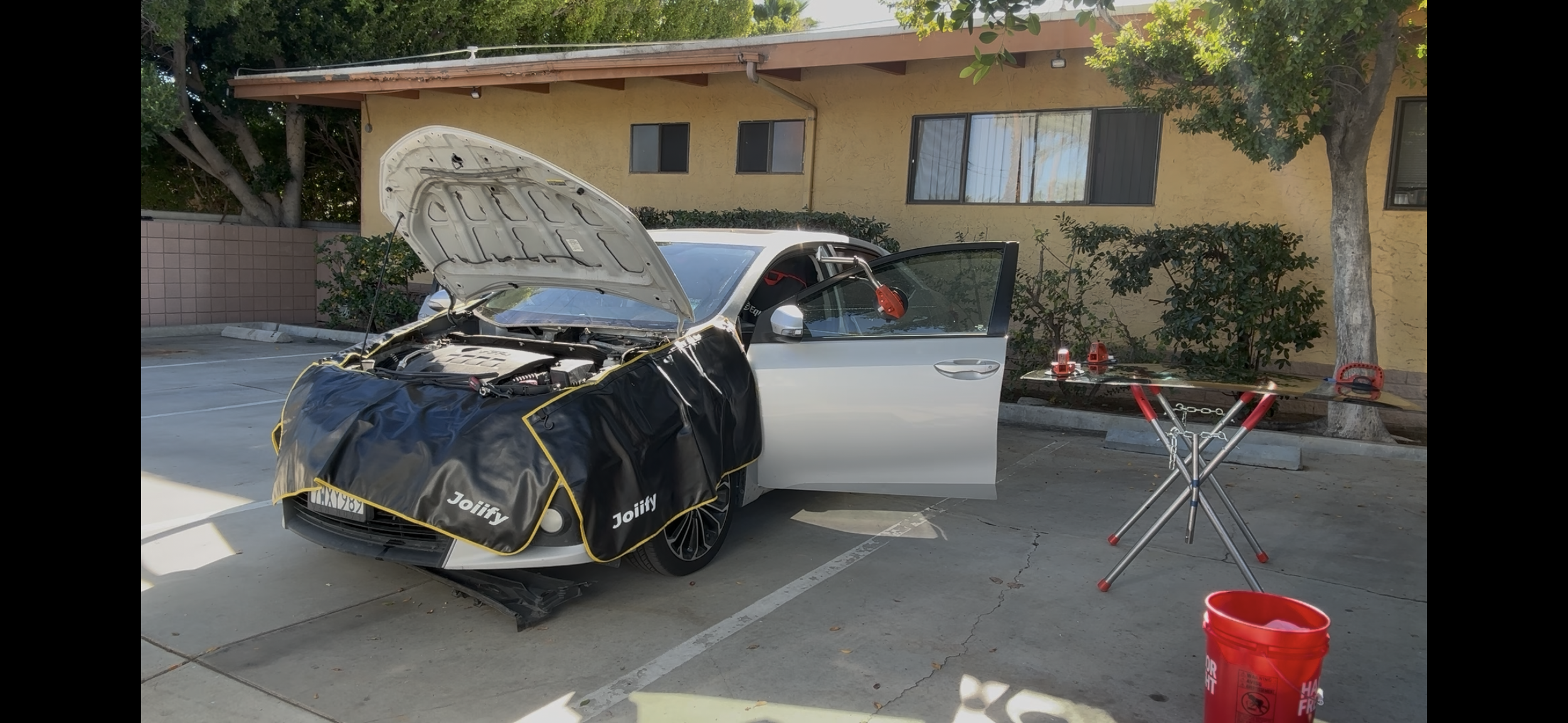 A silver car with its hood open and a black and yellow service cover over the front, parked in an outdoor parking lot next to a building with yellow walls and windows. There is a small table with red tools and equipment beside the car, including a ch