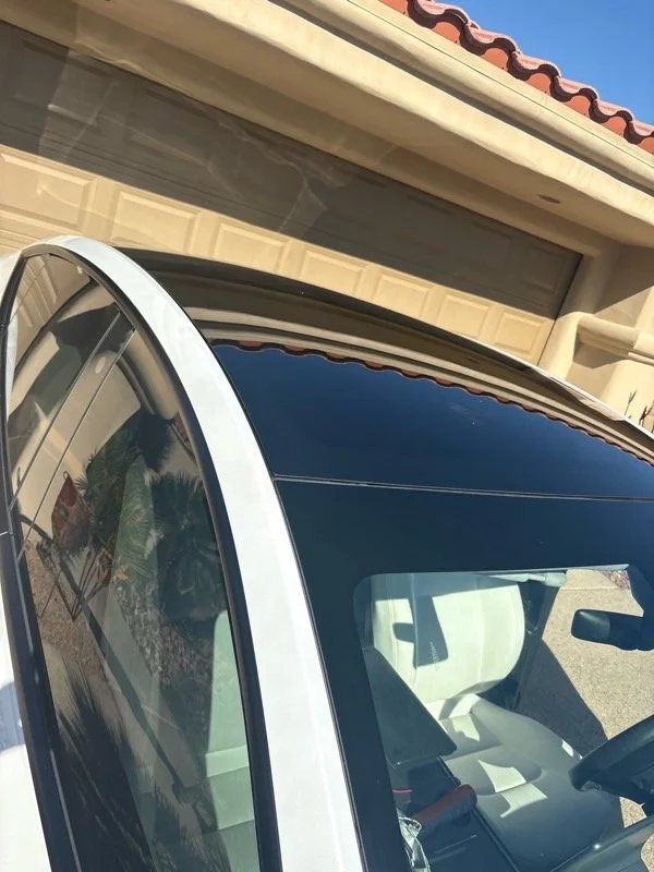 A white car parked outside a house with a beige garage door and a roof with red tiles. The windshield reflects palm trees.
