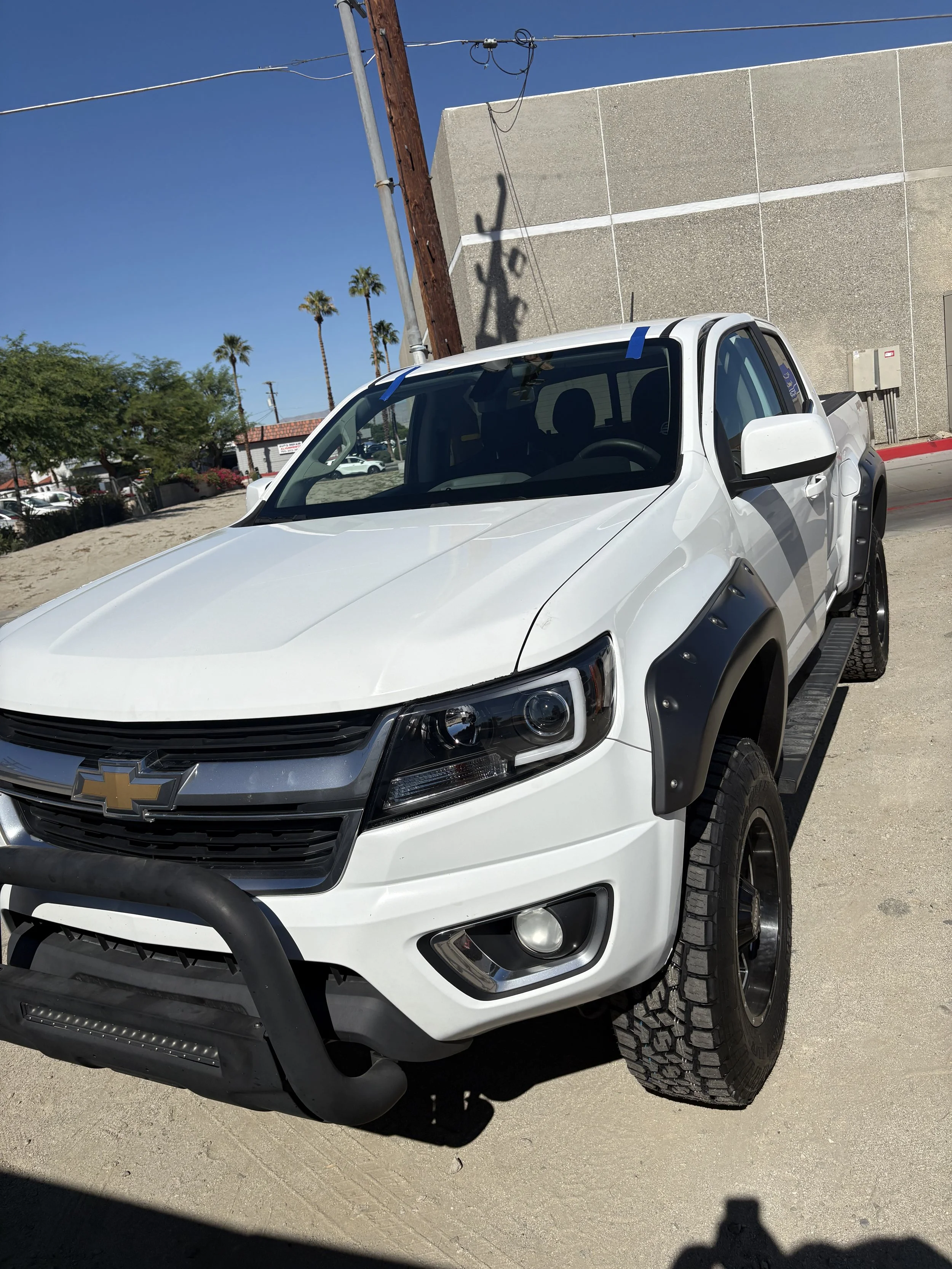 White pickup truck with black fender flares parked outdoors under a clear blue sky, with palm trees in the background.