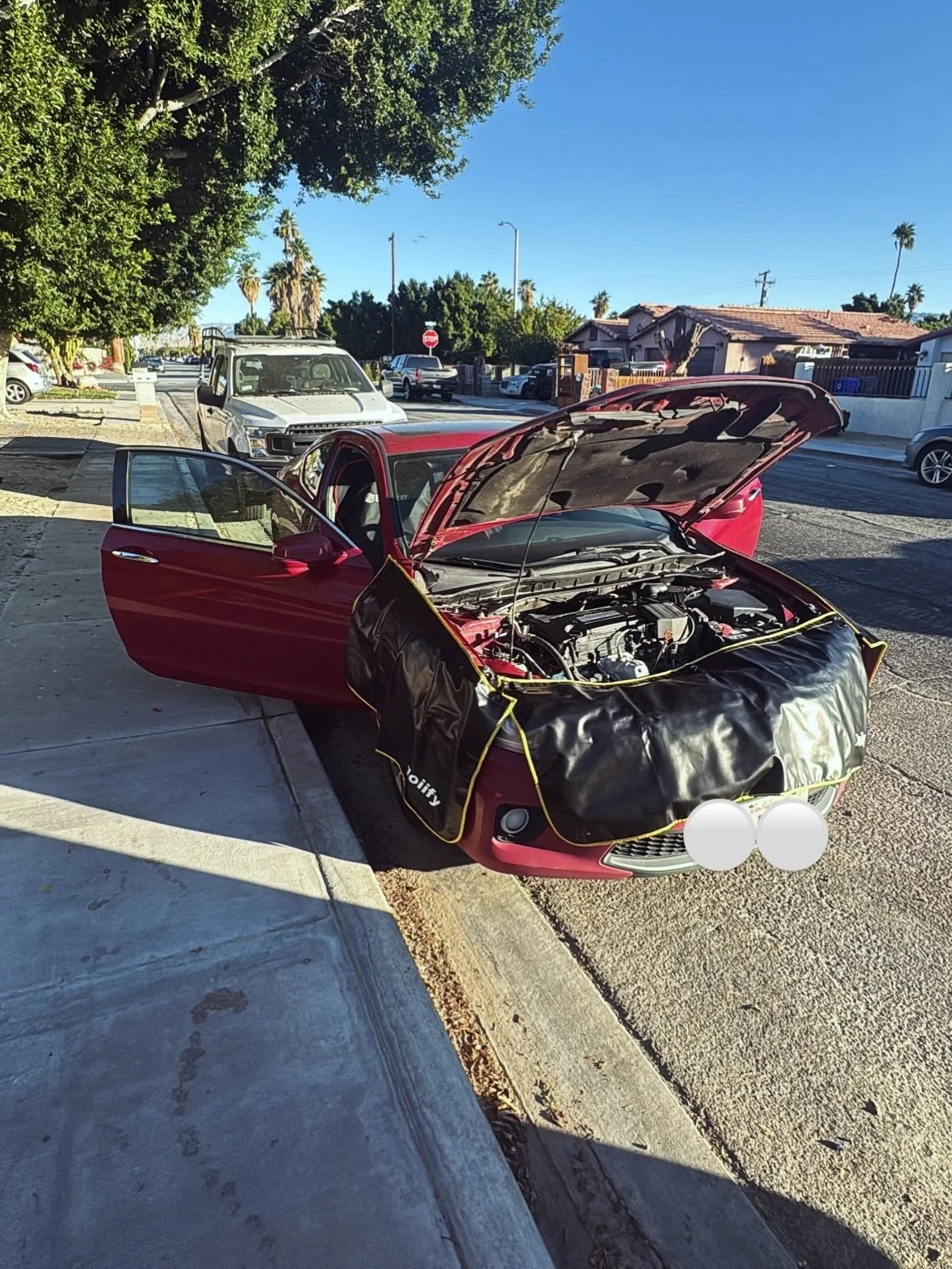 A red car with its hood open and a black cover on the front is parked on the side of the street in a residential neighborhood. The driver's door is open, and other cars are parked along the curb. There are trees, houses, and clear blue sky in the bac