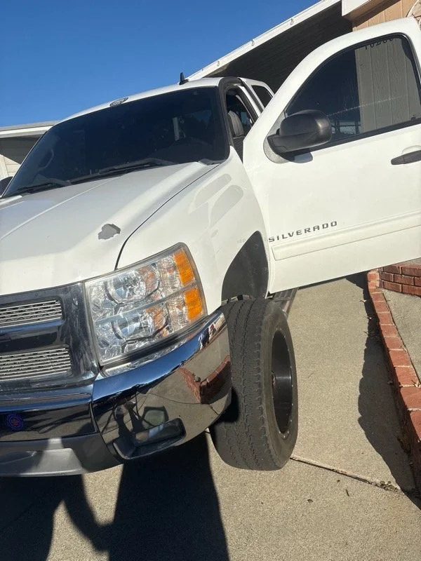 A white Chevrolet Silverado pickup truck with front wheels on the ground and rear wheels elevated on a block during daytime, parked next to a brick sidewalk.