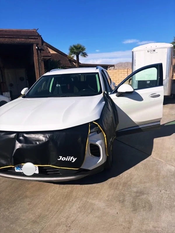 A white car with black car covers partially removed, parked on a driveway next to a house and a trailer. The car's front is uncovered, showing the logo 'Jolify' on the cover.