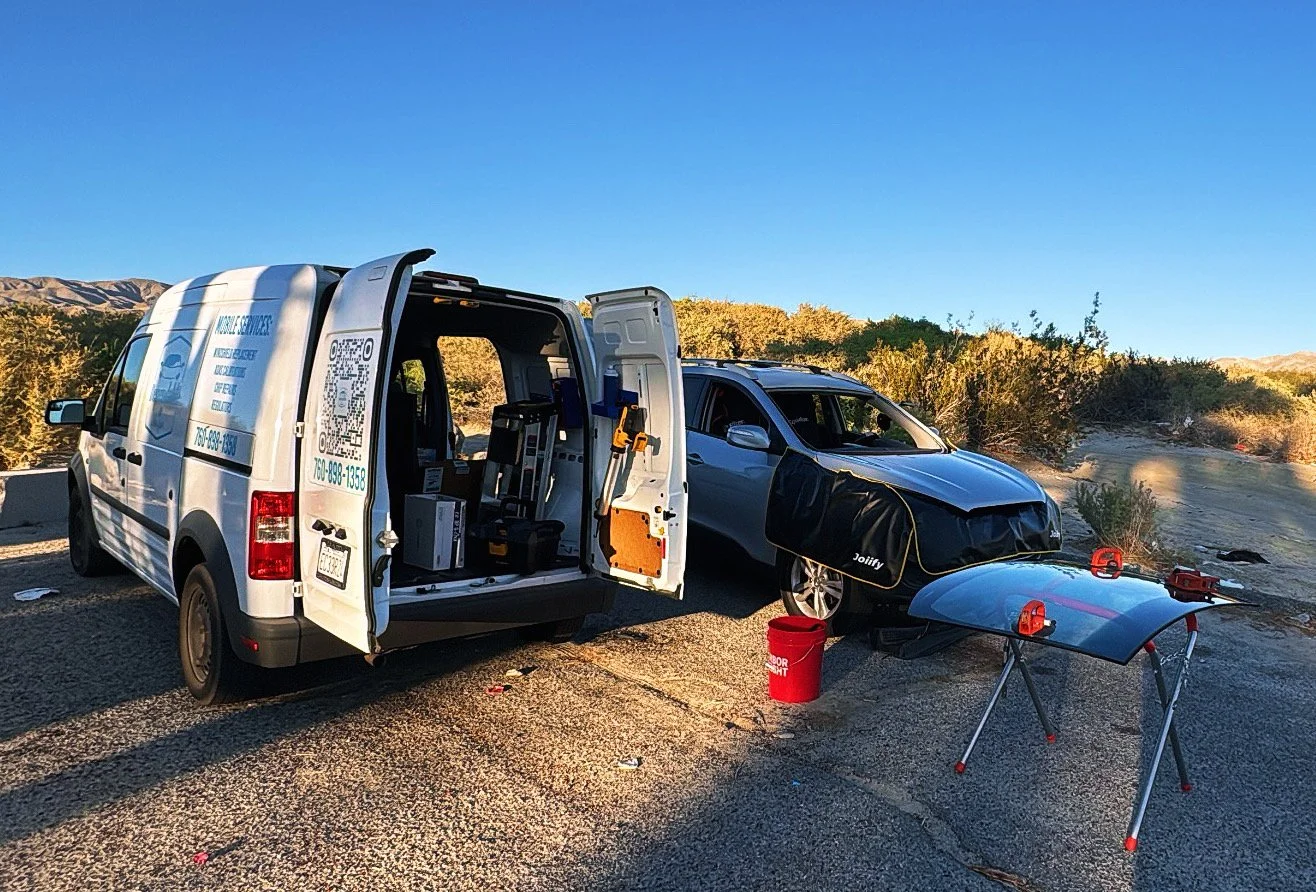 A vehicle repair service van parked on the side of a road with its side door open, next to a silver car with a missing front bumper, on a sunny day with desert landscape in the background. There is a red bucket, a car hood on a stand, and tools near 