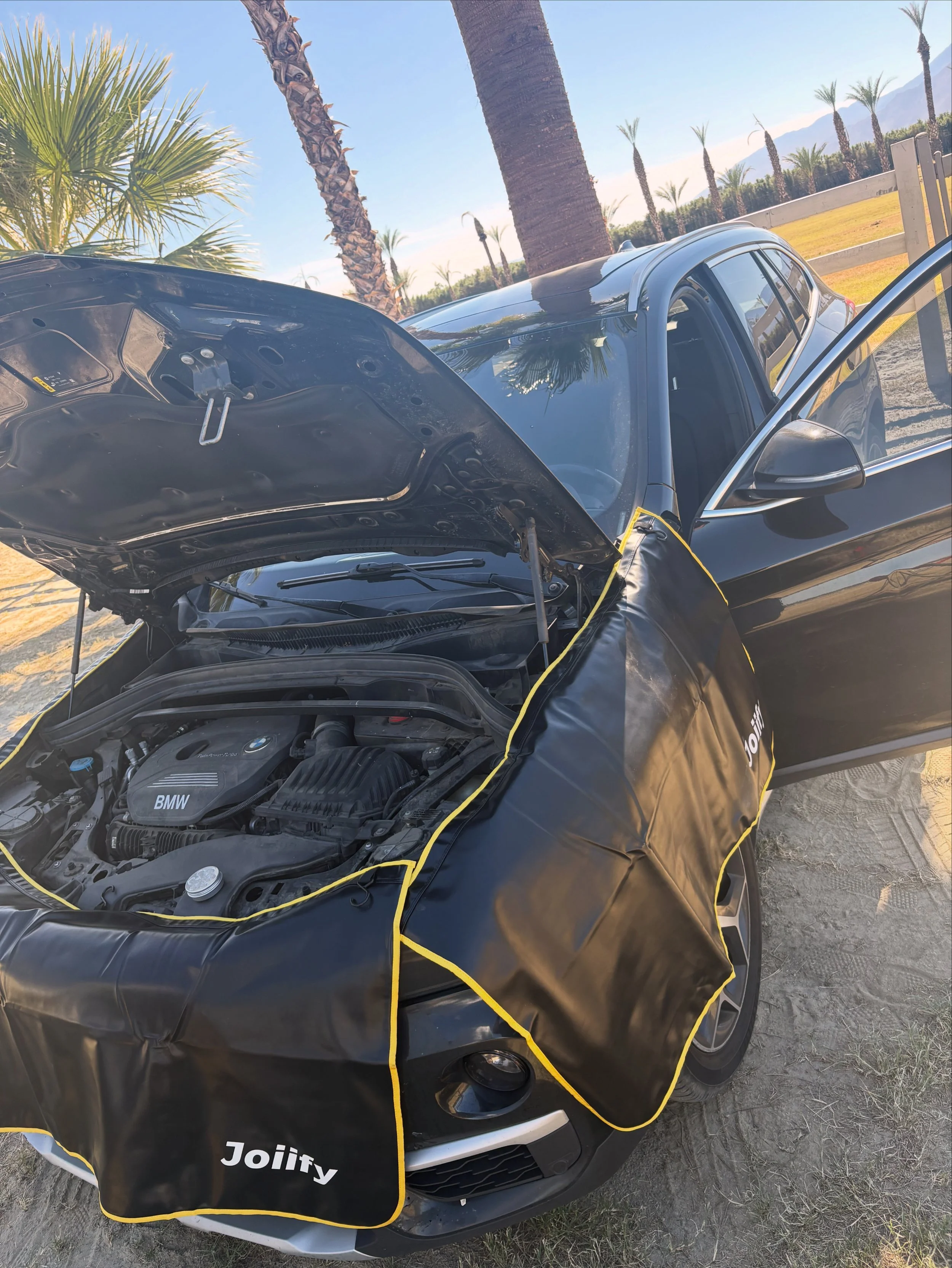 Black BMW car with the hood open, surrounded by a black sunshield labeled 'Joiify,' parked on sandy ground near palm trees and a fence, with mountains and blue sky in the background.