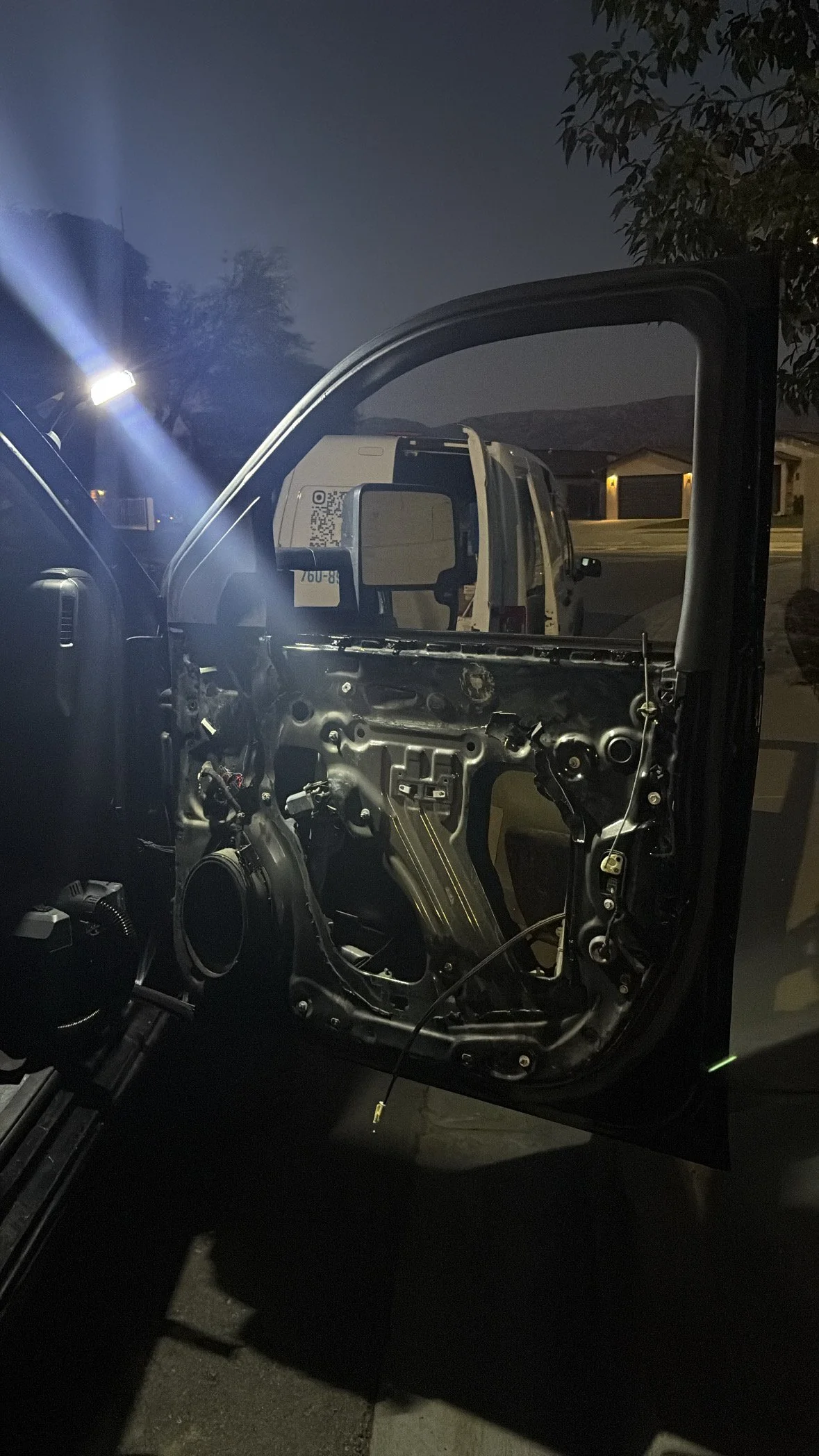 The interior of a vehicle with the door panel removed, exposing wiring and mechanical components, taken at night in a residential area with houses and a van visible in the background.
