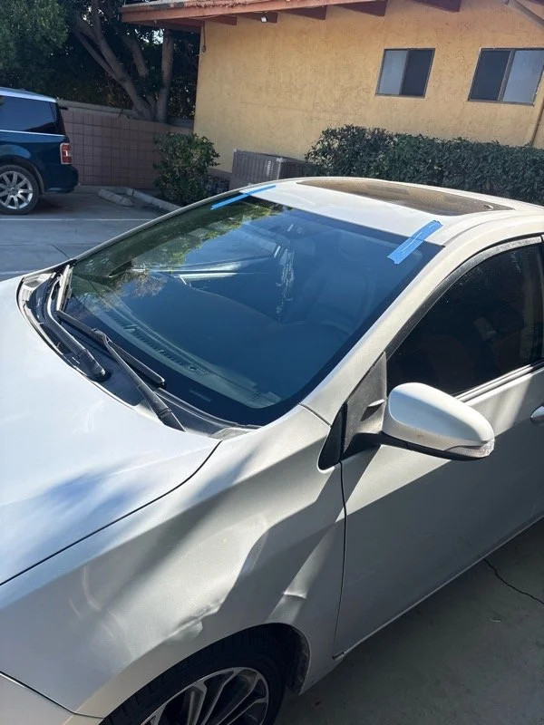 A white car parked in a driveway with tape on the windshield, indicating it may be new or for delivery, with residential buildings and other vehicles in the background.