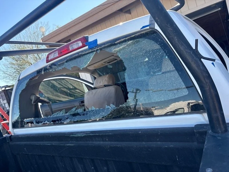 A pickup truck with a shattered rear window and cracked side window in a residential area with house and trees in the background.