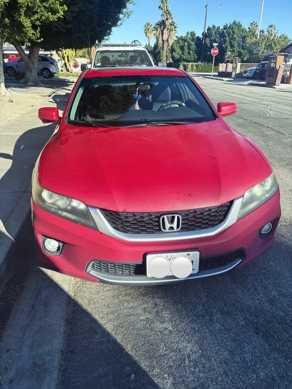 Red Honda car parked near the curb with a white vehicle behind it, in a sunny outdoor parking area with trees and a stop sign visible in the background.