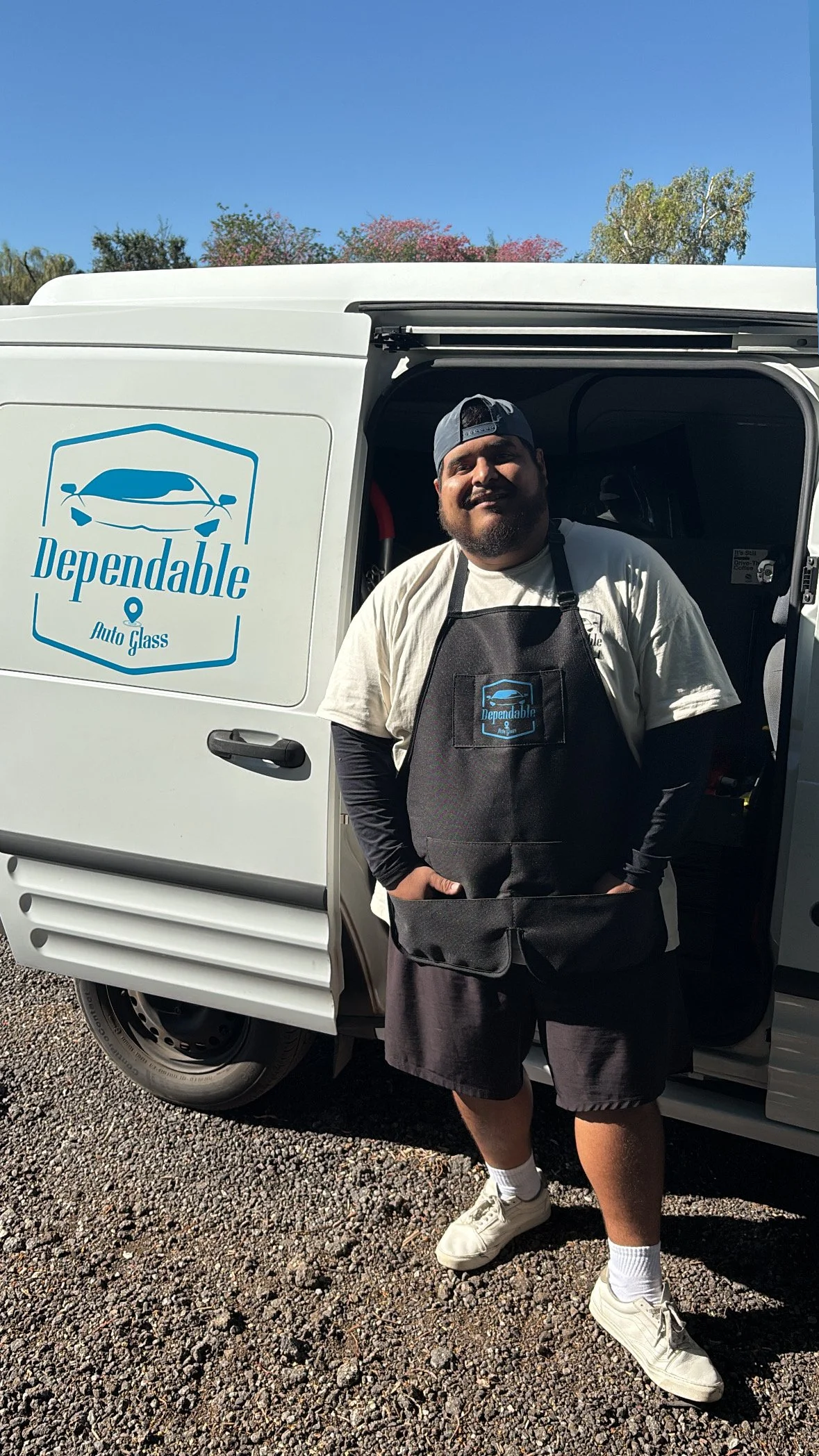 A man standing in front of a white service van with 'Dependable Auto Glass' logo. The man wears a black apron with the same logo, a white t-shirt under a long-sleeved black shirt, shorts, white sneakers, and a backwards cap. The background shows a clear blue sky and some trees.