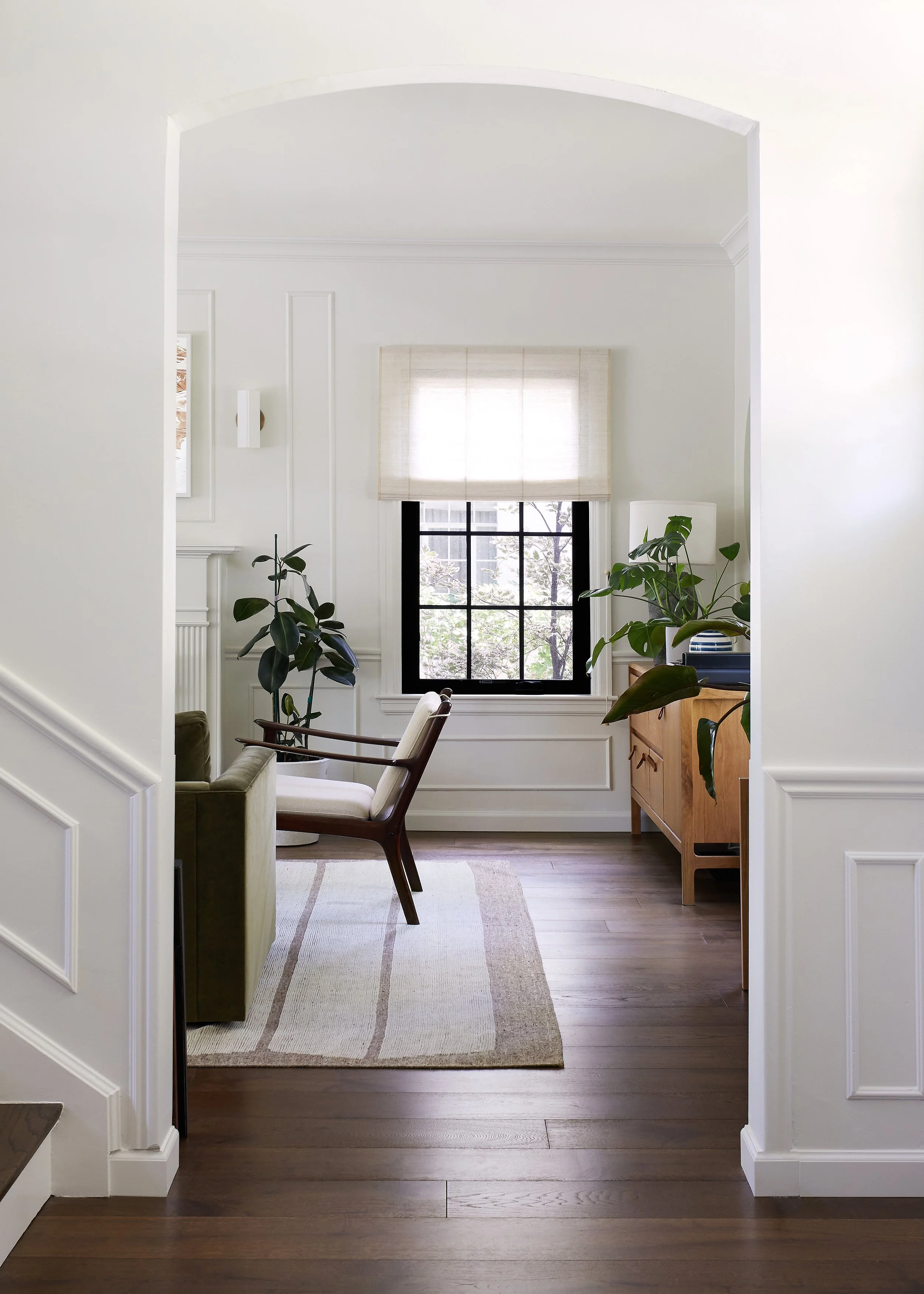 Living room with white walls, wooden flooring, a large window with a beige shade, green plants, a wooden cabinet, and a vintage armchair.