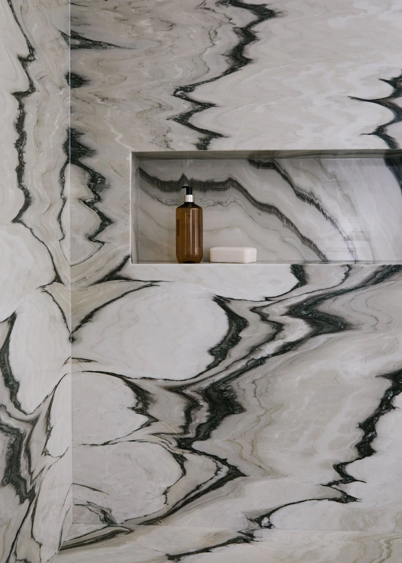 Marble shower with a built-in shelf holding a brown bottle and a bar of soap.