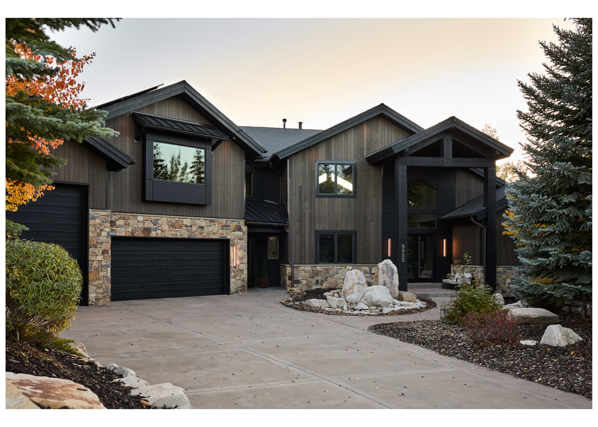 Modern house with dark wood siding, stone accents, and black garage doors, surrounded by trees and landscaped yard at sunset.