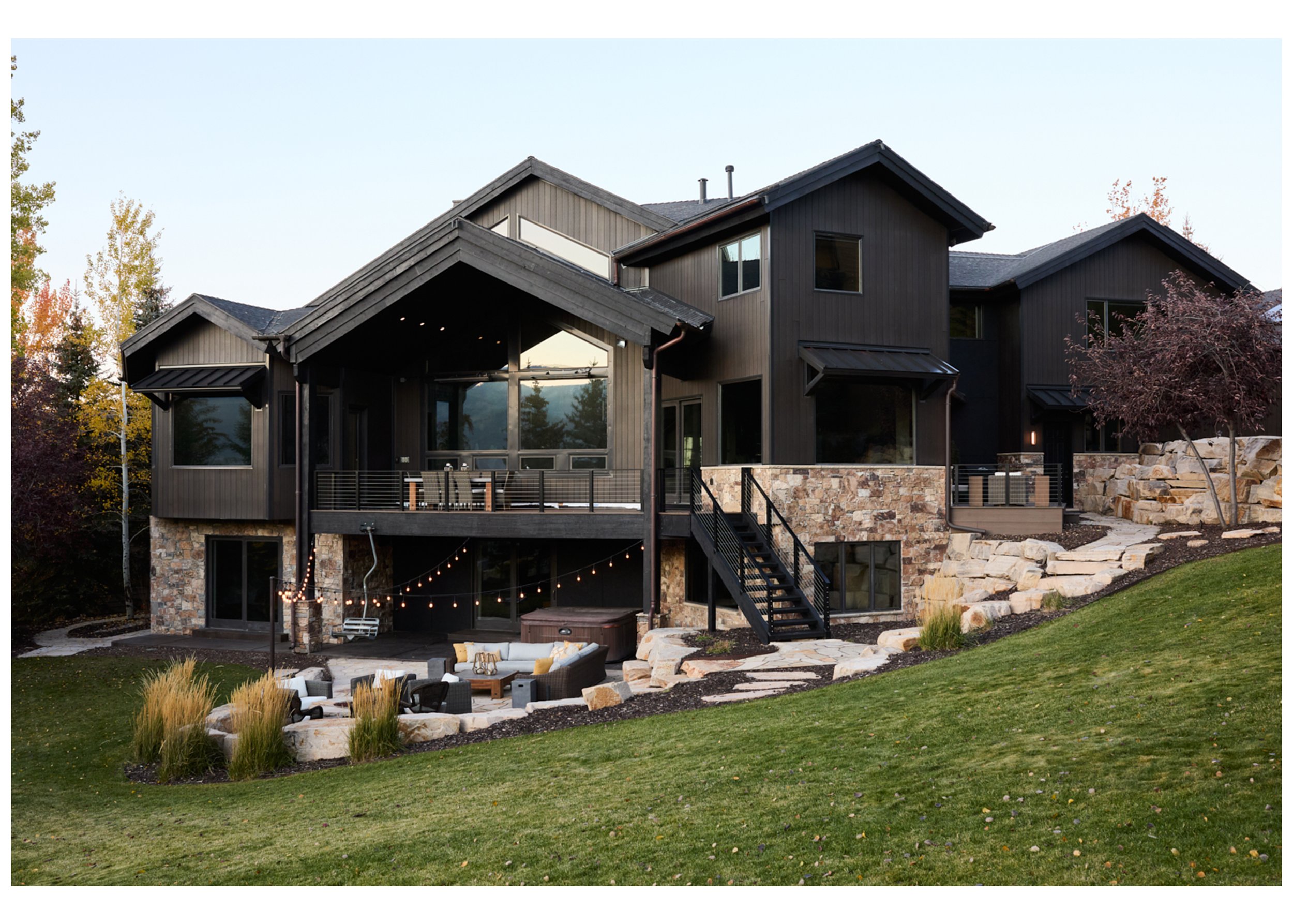 Modern multi-level house with dark wood siding, stone accents, and large windows, featuring an outdoor patio with seating, string lights, and landscaped yard with grass and rocks.