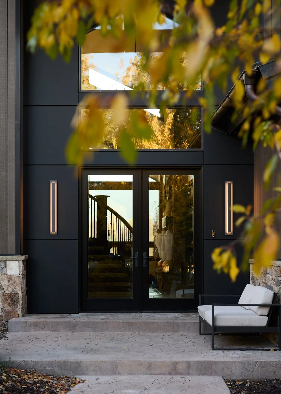 Modern house entrance with black double glass doors, stone steps, and a white outdoor bench, surrounded by trees with autumn leaves.