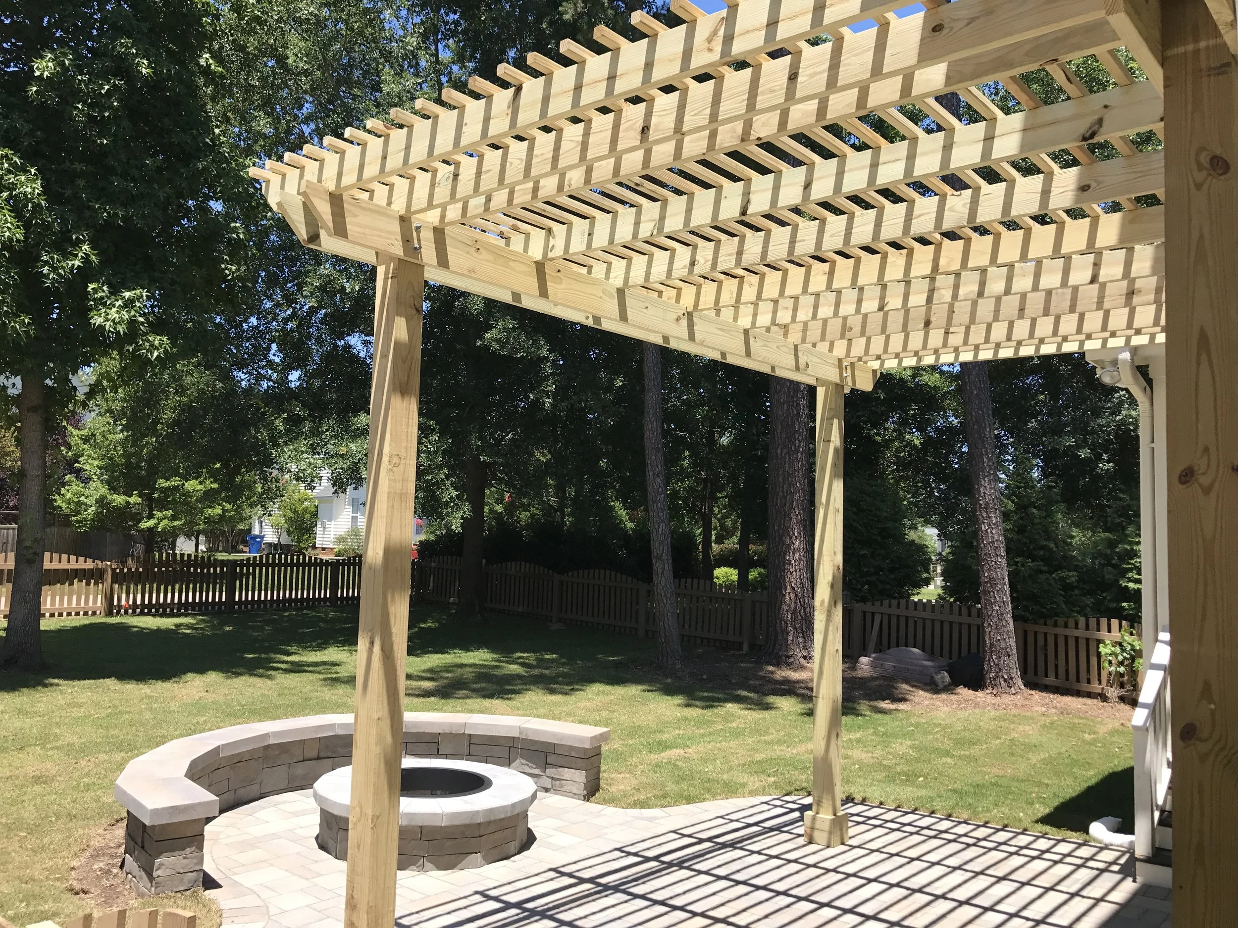 A wooden pergola with lattice in a backyard paver fire pit on paver patio with matching paver seat wall surrounded by mature trees.
