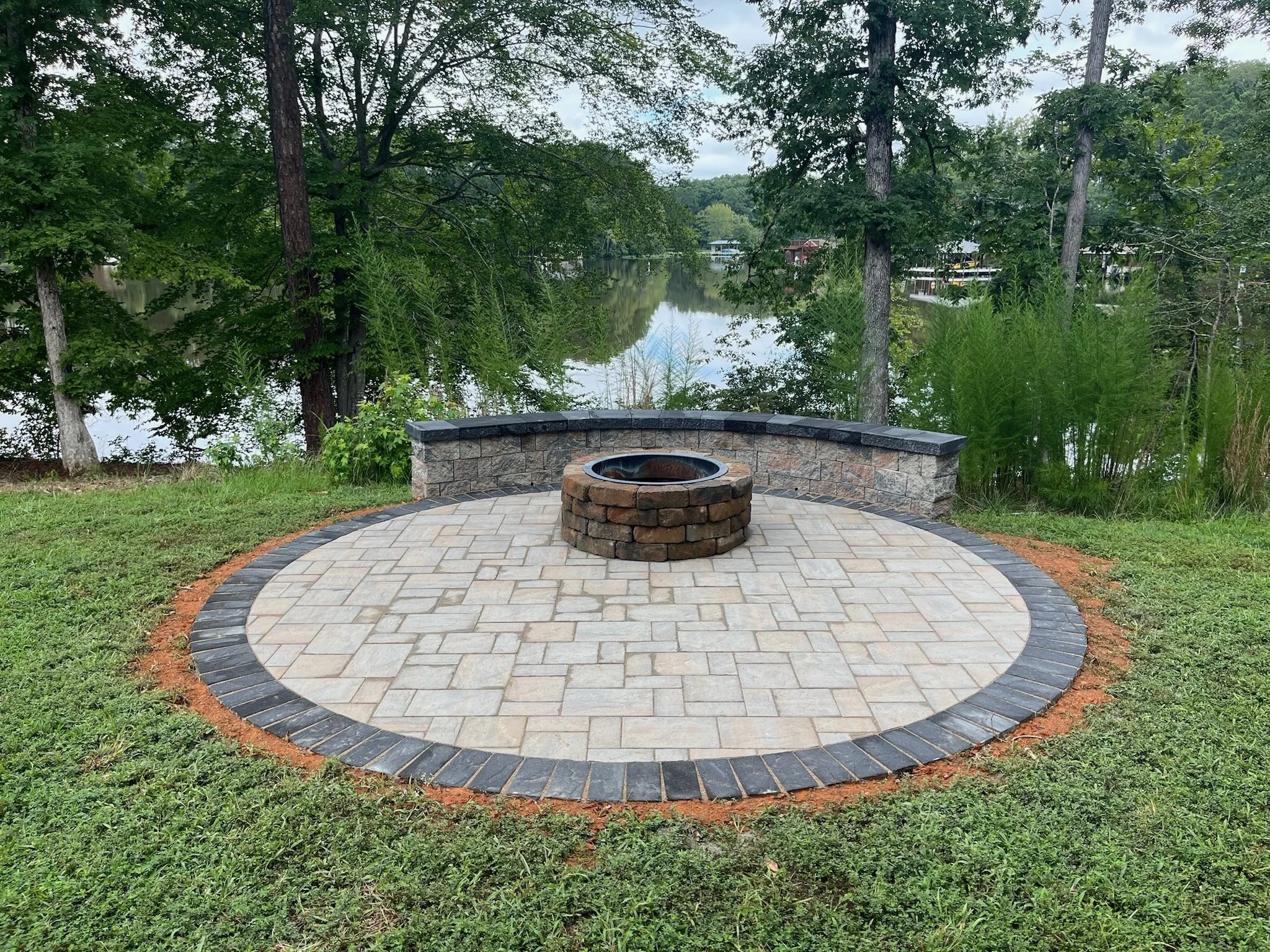 Paved circular paver patio area with a paver fire pit at the center, surrounded by a paver seat wall, in a grassy backyard with trees and a lake in the background.