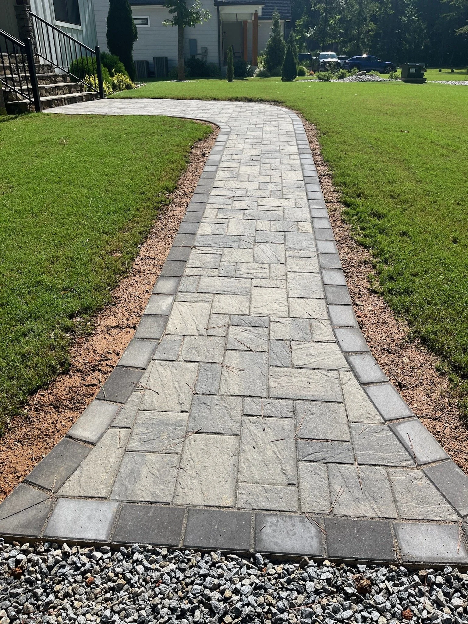 A curved paver walkway with a pattern of rectangular pavers bordered by darker paver stones, leading to a grassy yard.