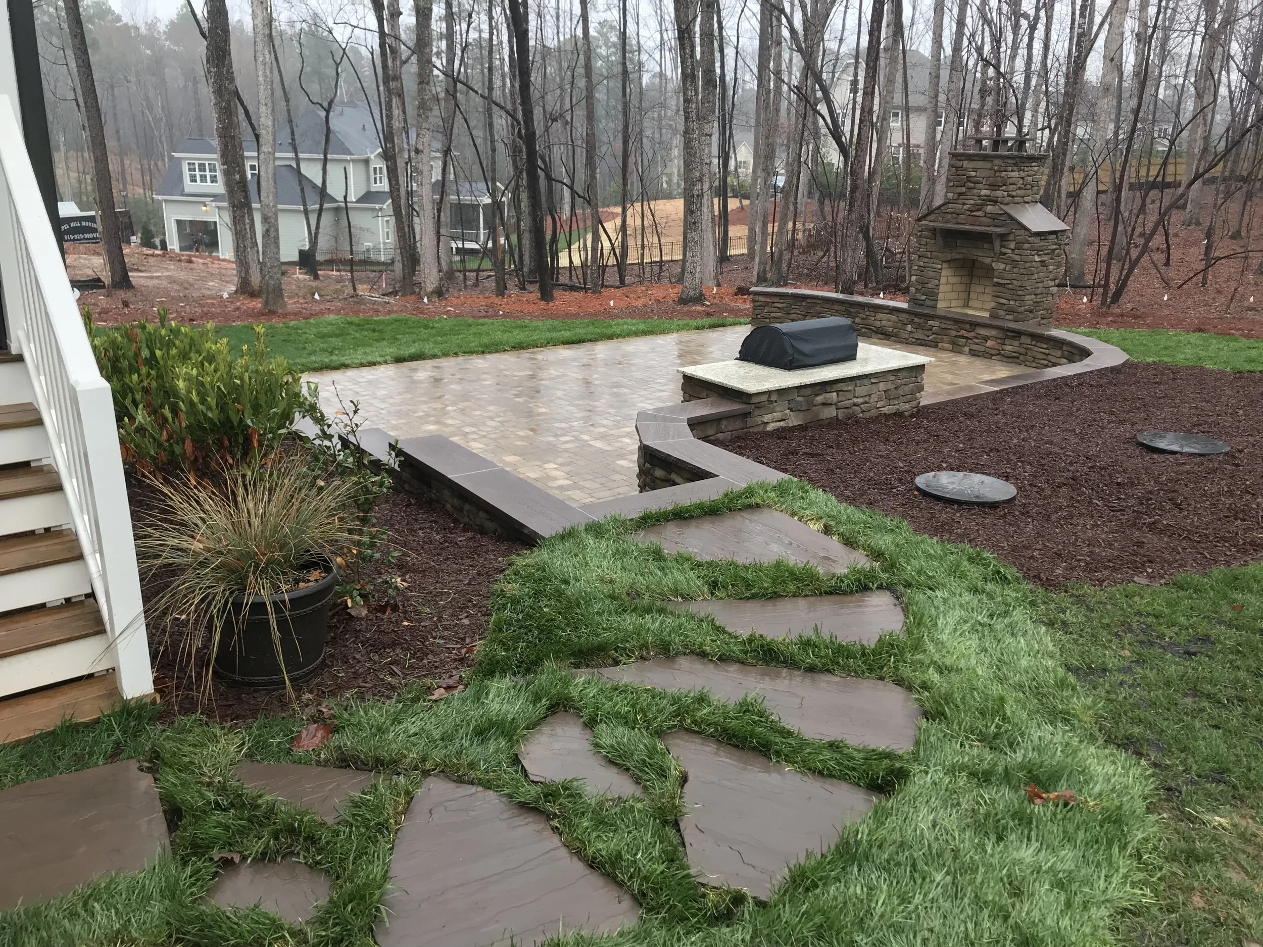 Backyard with flagstone walkway, paver fire pit area and retaining wall with paver stairs leading to house, surrounded by trees and grass.