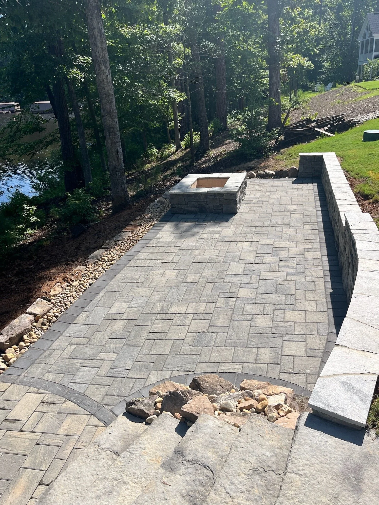 A newly paved stone patio with a fire pit at the center, bordered by a stone retaining wall, with a wooded area and water in the background.