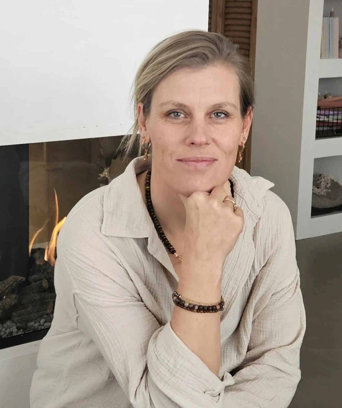 Portret van een vrouw met blond haar, zittend voor een haardvuur, in een huis met boekenplank op de achtergrond.