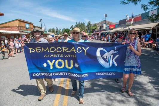 People holding a blue Democratic rally banner at a parade in Okanogan County, with spectators lining the street.