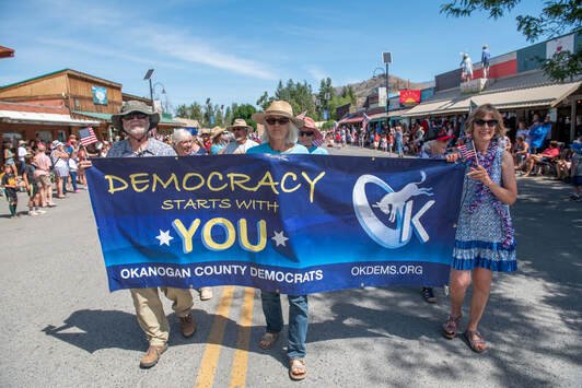 Three people holding a blue Democratic parade banner that says "Democracy starts with you" at a street event with people and shops in the background.