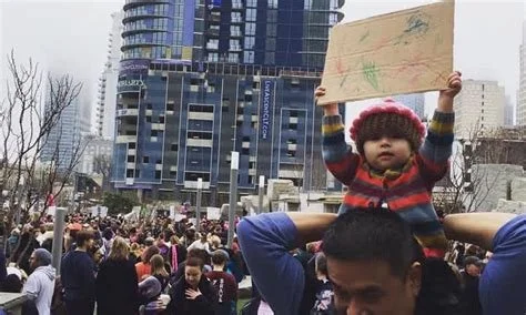 A young child seated on an adult's shoulders holding a cardboard sign in a busy outdoor setting with a crowd and tall modern buildings in the background.