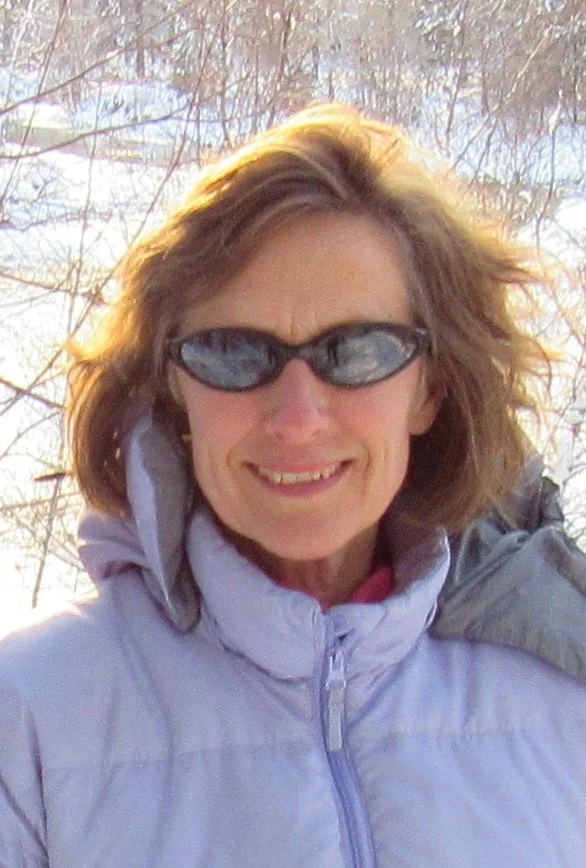 Picture of Halley Hart wearing sunglasses and a light gray jacket, standing outdoors with bare trees and snow in the background.