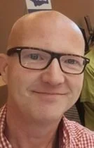 Close-up of a smiling man with glasses, bald head, and wearing a red checkered shirt.