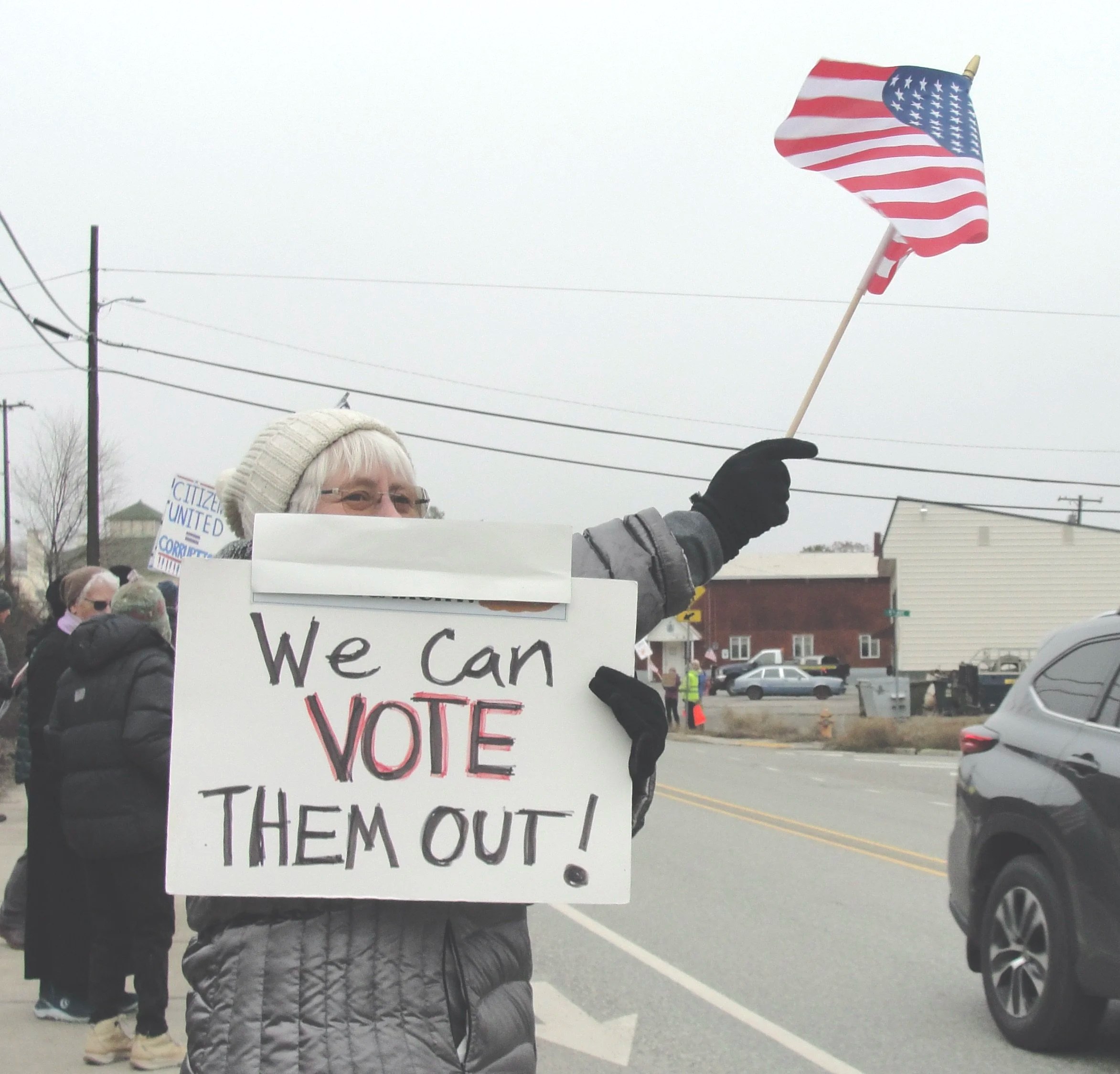 A protester on the street, holding a sign that says 'We can VOTE them out!' and waving an American flag.